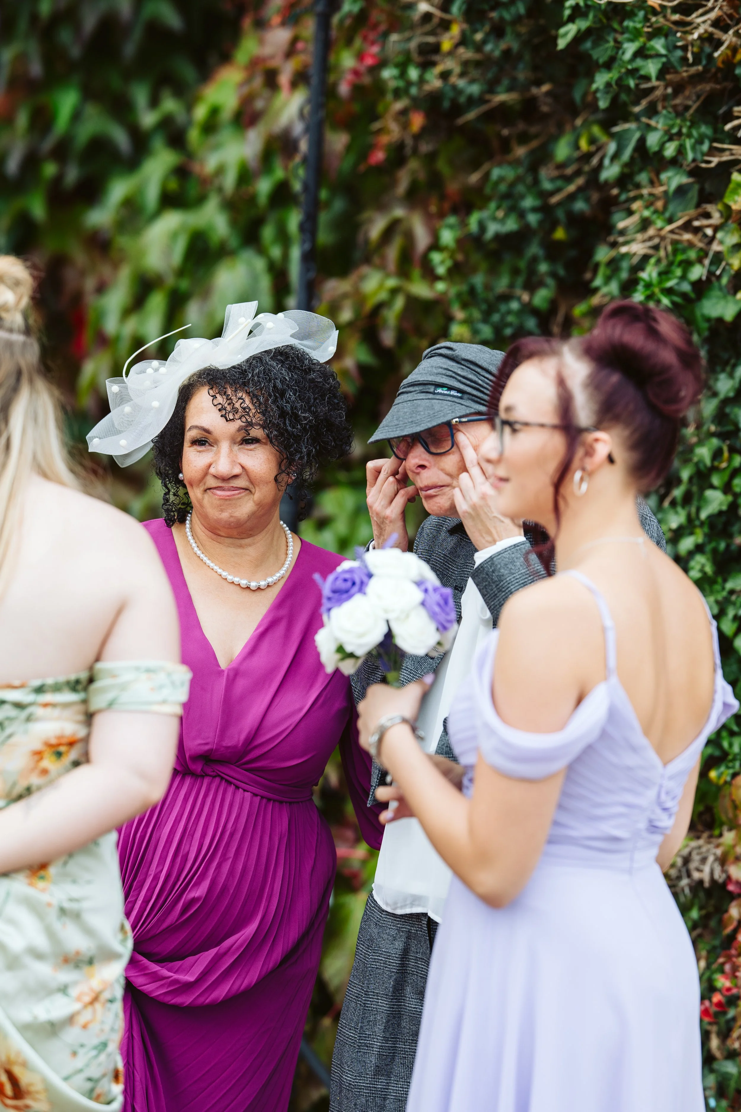 A group of women at a wedding, with two women clearly visible, one wearing a purple dress and a decorative white hat, and the other wearing a light lavender dress and glasses, holding a bouquet of white and purple flowers, with a lush green hedge in the background.