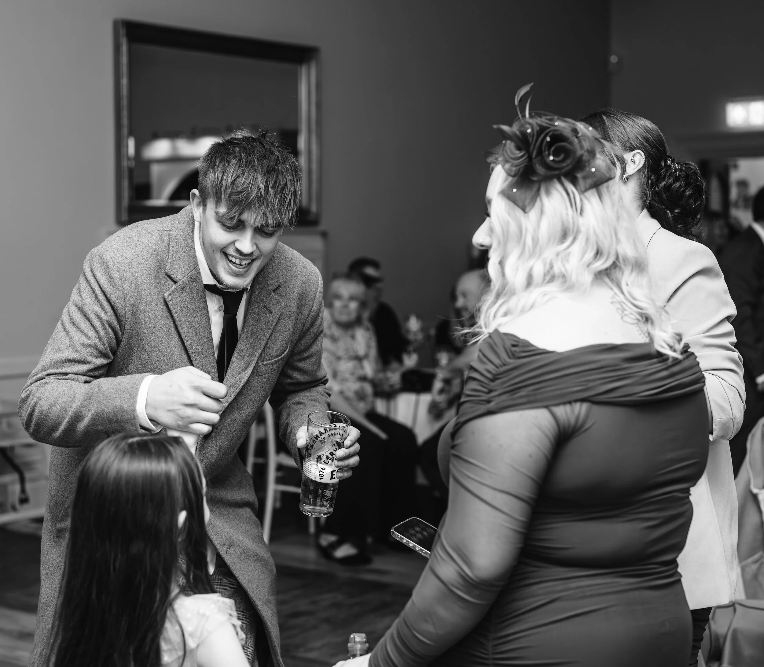 A young man in a suit and a woman with a fascinator hat are smiling and talking to a girl sitting in front of them at a social gathering or wedding reception, with guests seated at tables in the background.