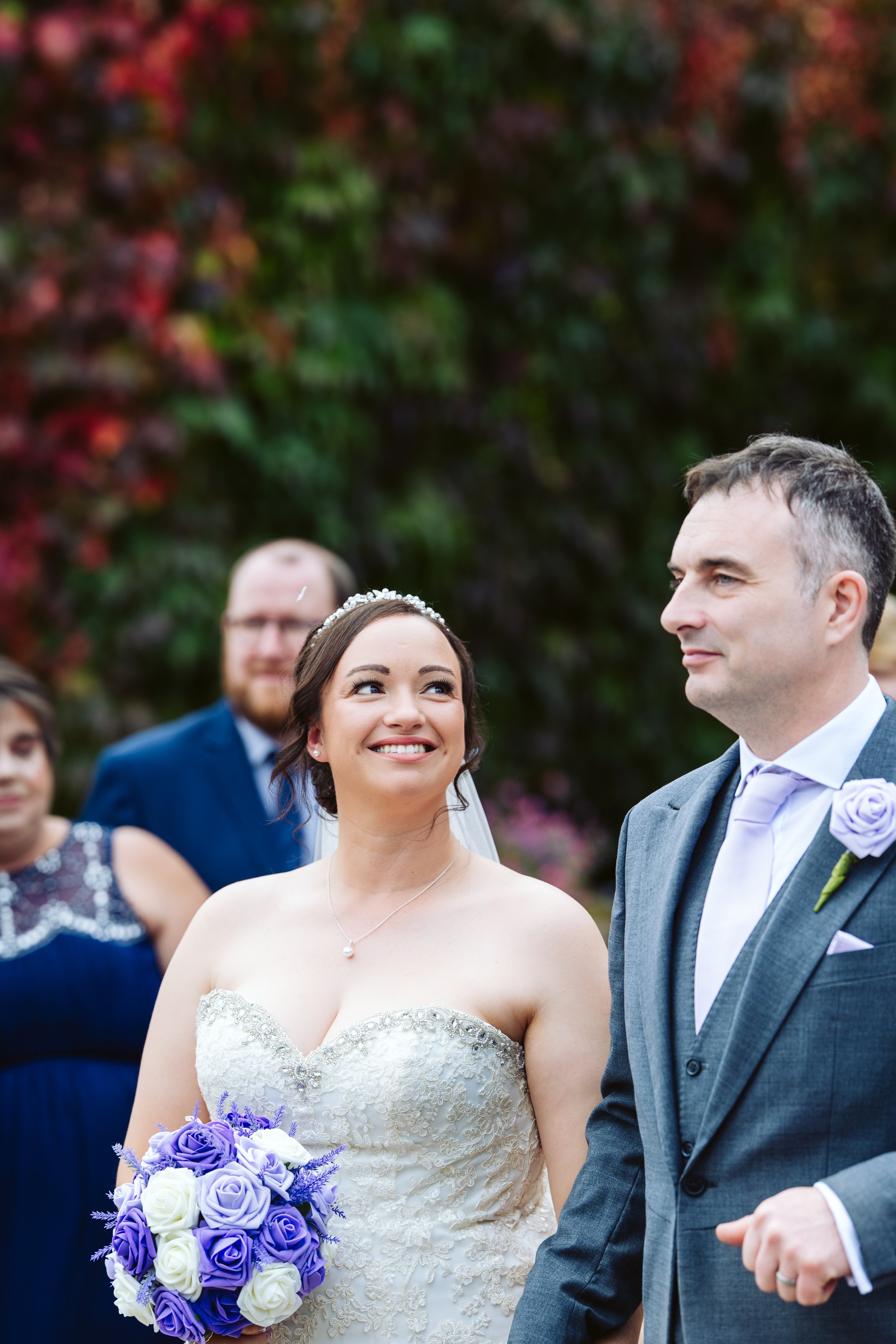 A bride in a strapless wedding dress holding a bouquet of purple and white roses looks at her groom. The groom wears a gray suit with a lavender tie and boutonniere. In the background, friends or family members are blurred, with autumn foliage behind them.