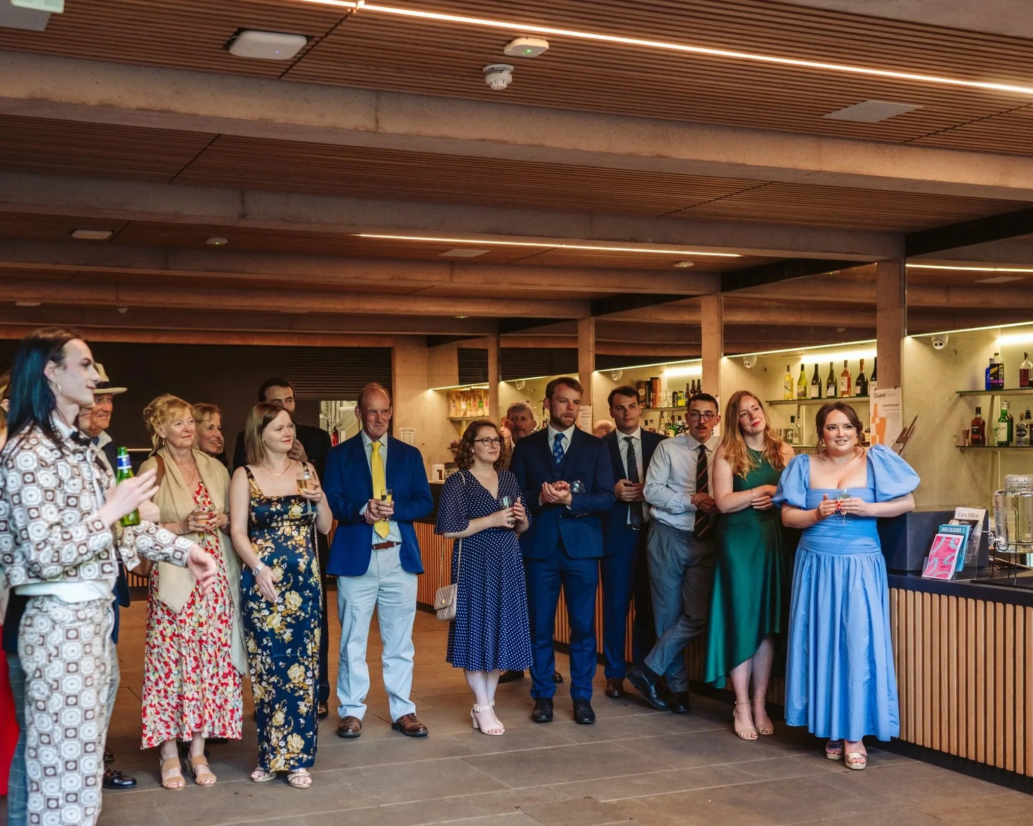 A group of formally dressed people gathered in a modern bar or lounge, listening attentively to someone outside the frame, with shelves of bottles visible in the background.