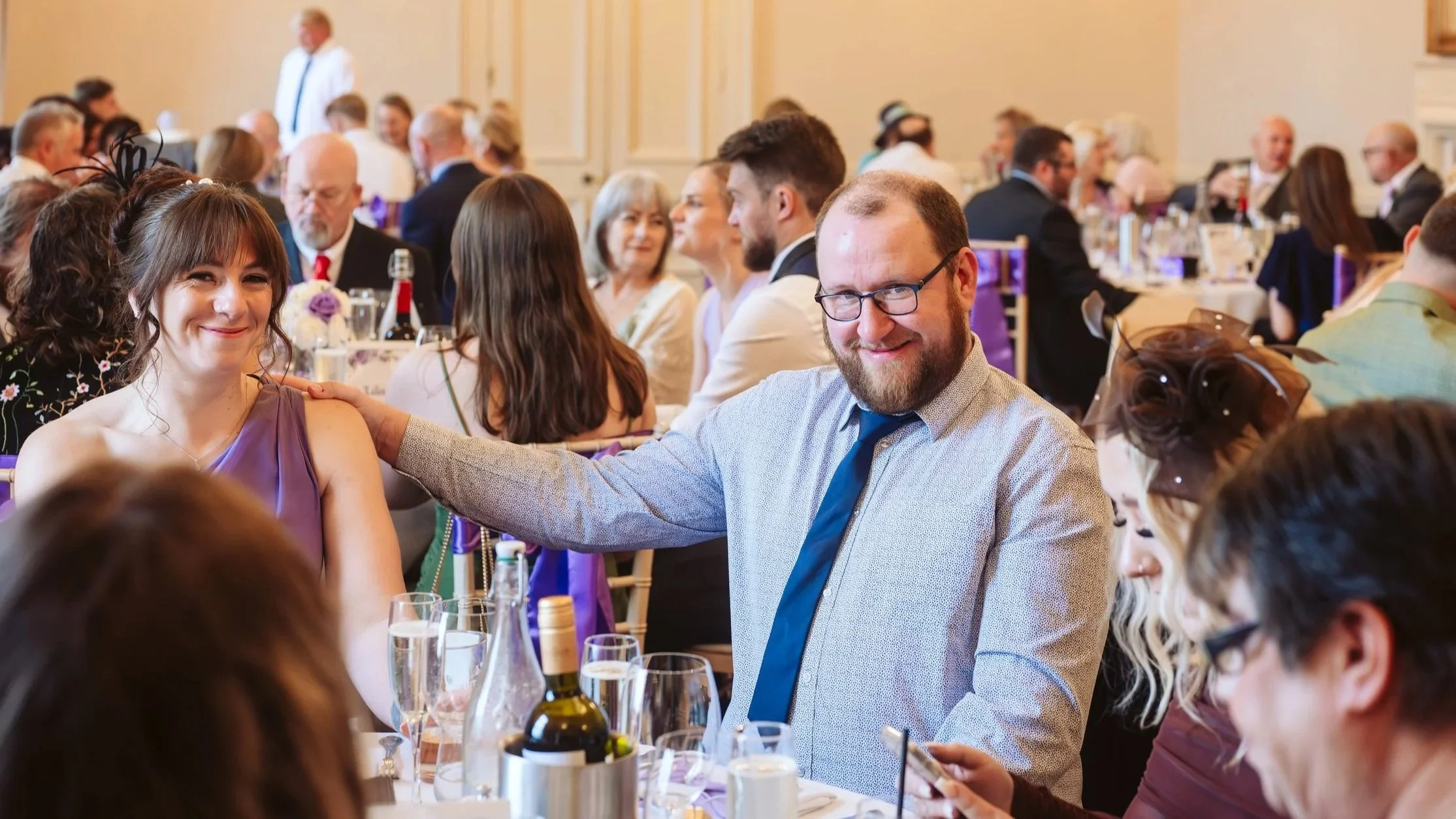 People gathered at a formal event, sitting at decorated tables with drinks, in a well-lit banquet hall.