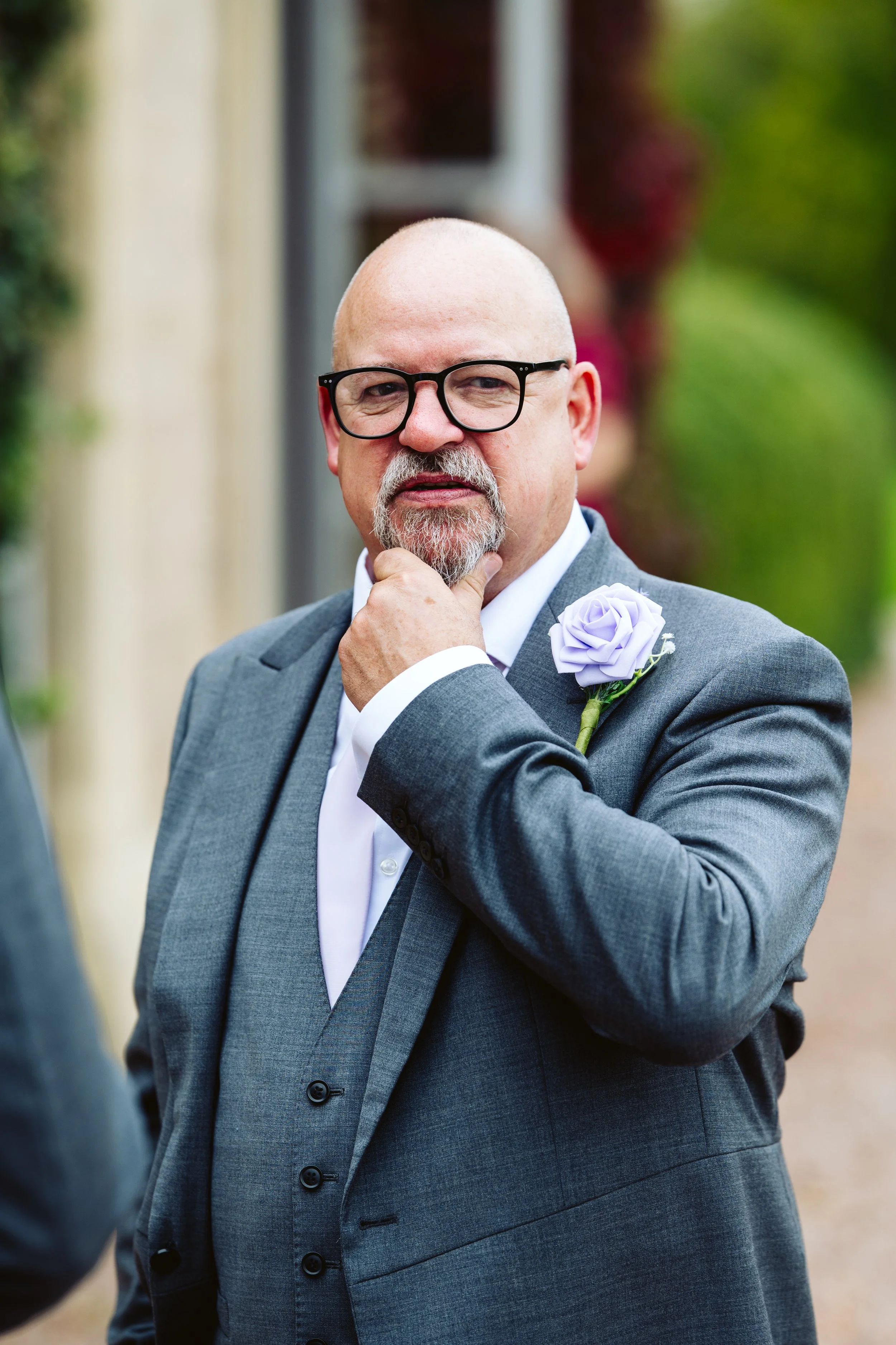 A man with glasses and a beard, dressed in a gray suit with a white shirt, has a thoughtful expression and touches his chin, standing outdoors with blurred greenery in the background.