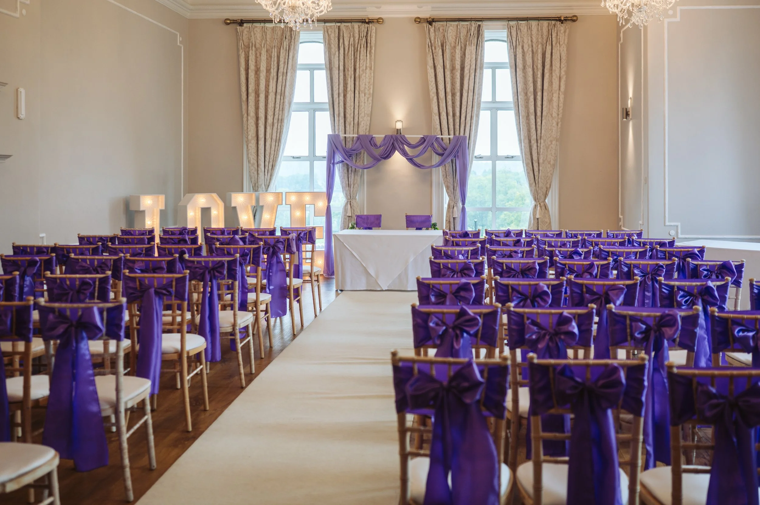 Wedding ceremony setup in a bright room with large windows and draped curtains, decorated with purple bows on chairs, illuminated 'LOVE' sign near the altar, and an elegant table at the front.