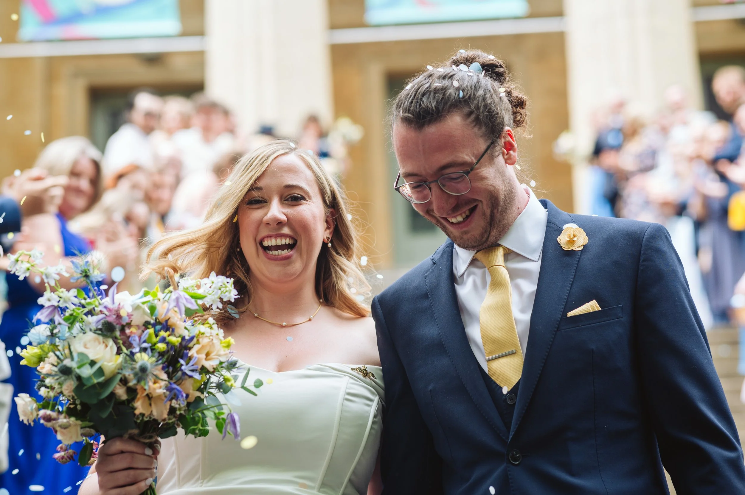 A bride and groom smiling and celebrating, with the bride holding a bouquet of flowers, surrounded by guests at a wedding reception.