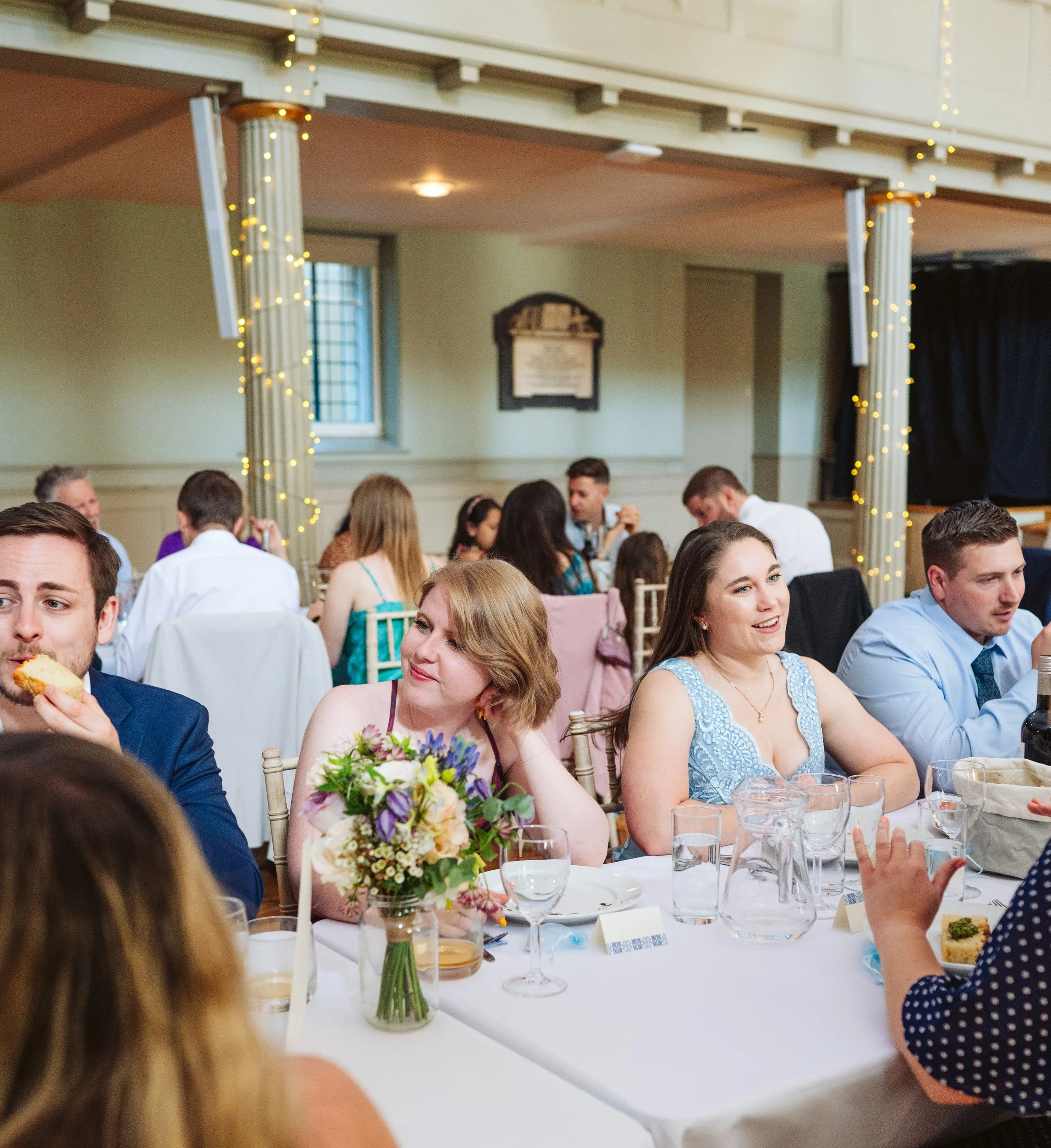 People seated at a decorated banquet table during a celebration or wedding reception, with floral arrangements, glasses, and tableware, in a well-lit room with string lights and columns.