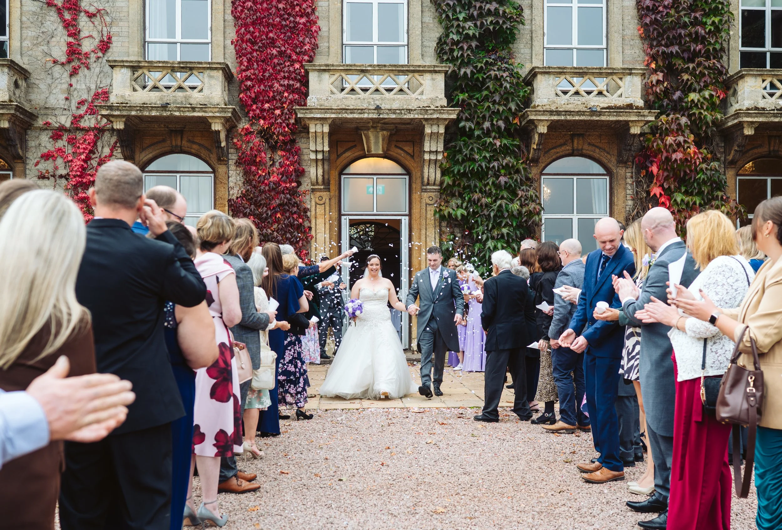 Wedding celebration with a bride and groom walking out of a building while guests throw confetti, surrounded by a crowd of guests clapping and taking photos, with a stone building decorated with red and green ivy in the background.