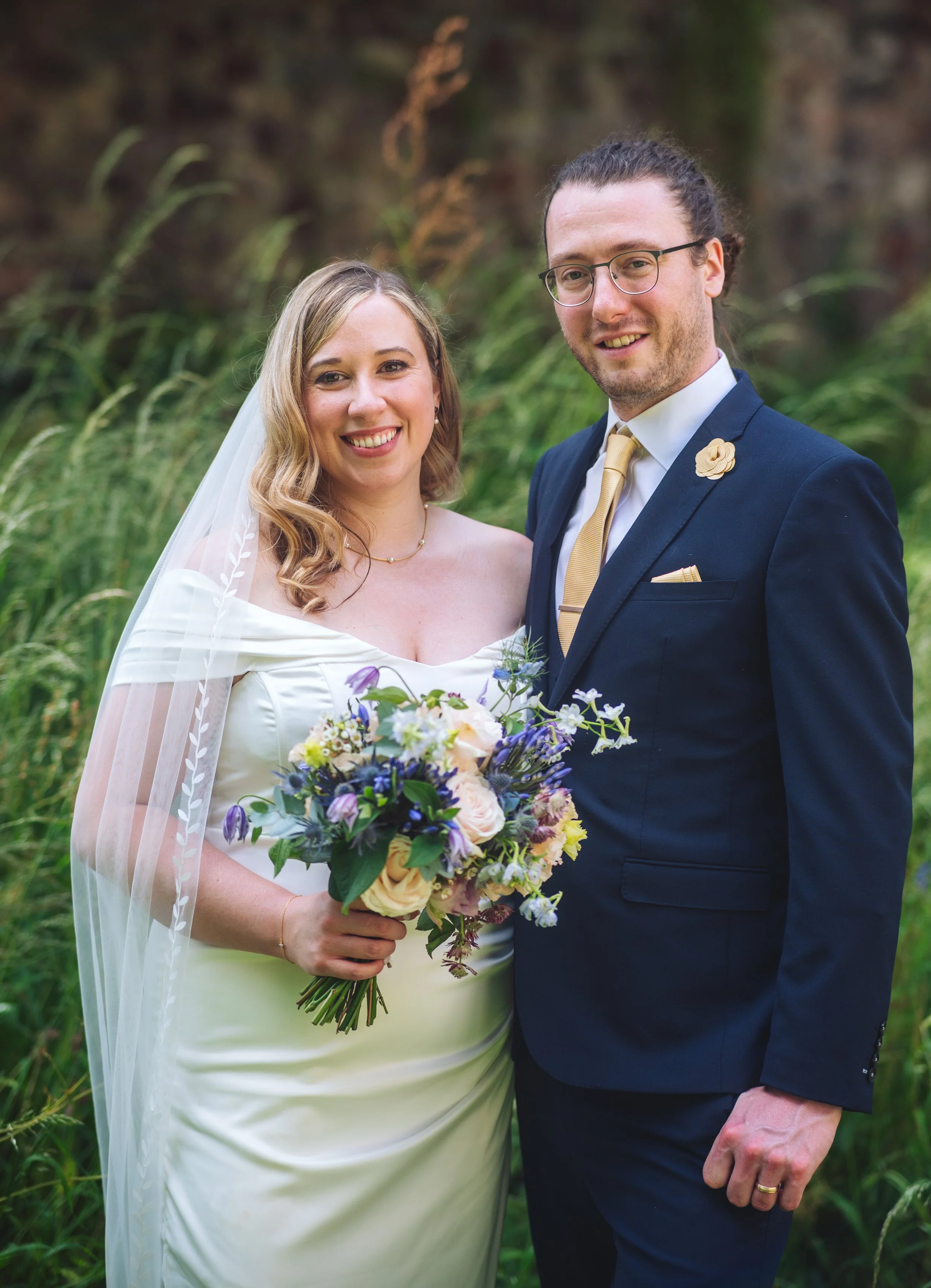 A bride and groom smiling outdoors during their wedding, with the bride holding a bouquet of flowers and wearing a white dress with a veil, and the groom dressed in a navy suit with a gold tie and boutonniere, standing in front of green foliage.