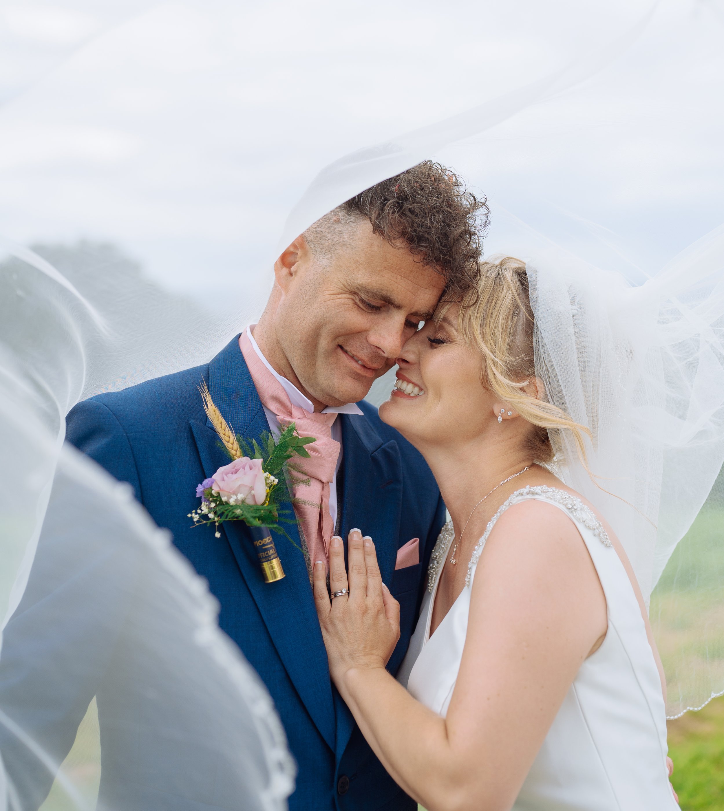 A bride and groom sharing a romantic moment on their wedding day under a veil outdoors, smiling and leaning their foreheads together.