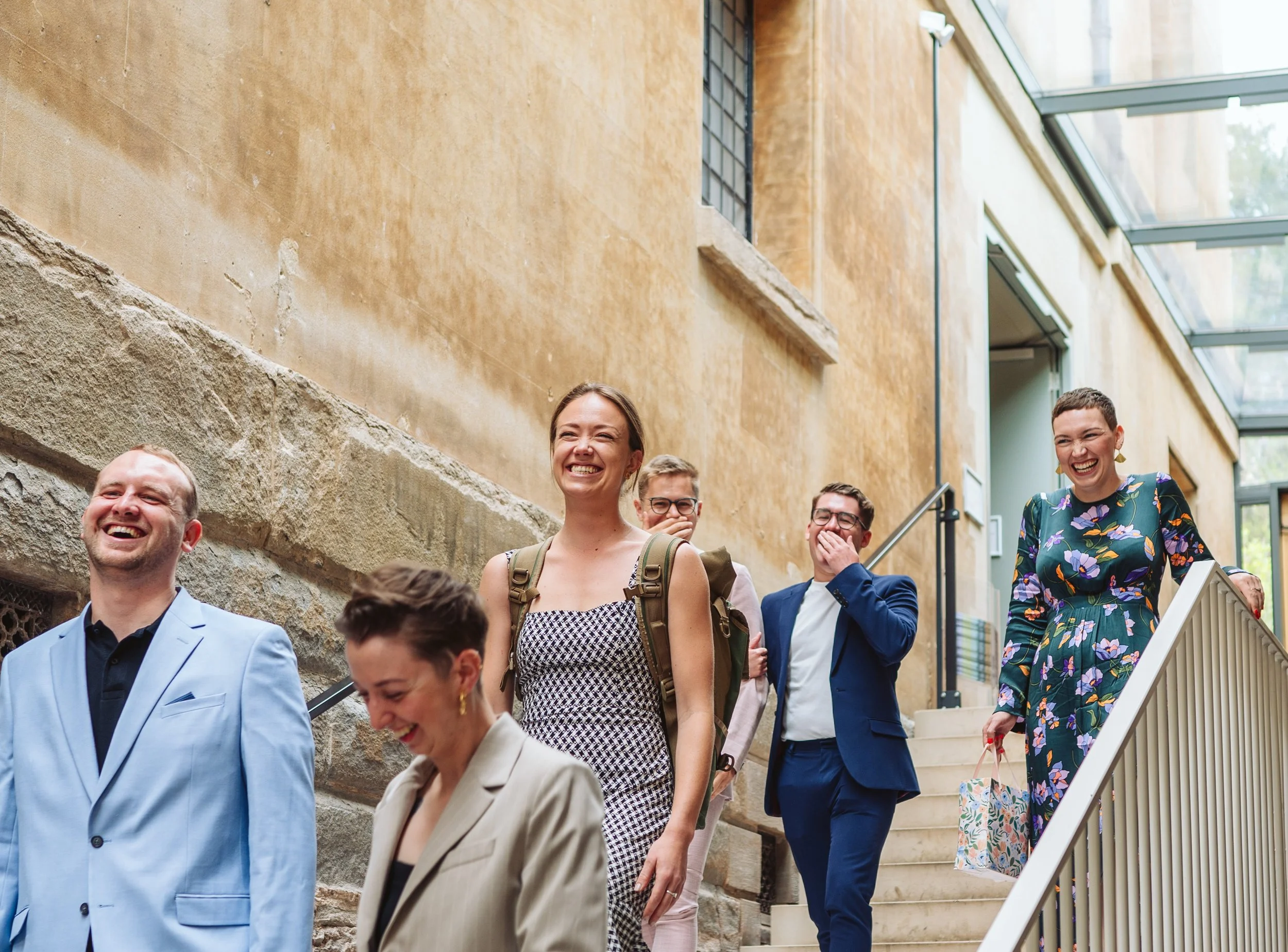 Group of six diverse professionally dressed people walking down a staircase, smiling and laughing in a well-lit, modern indoor space with beige walls and large windows.
