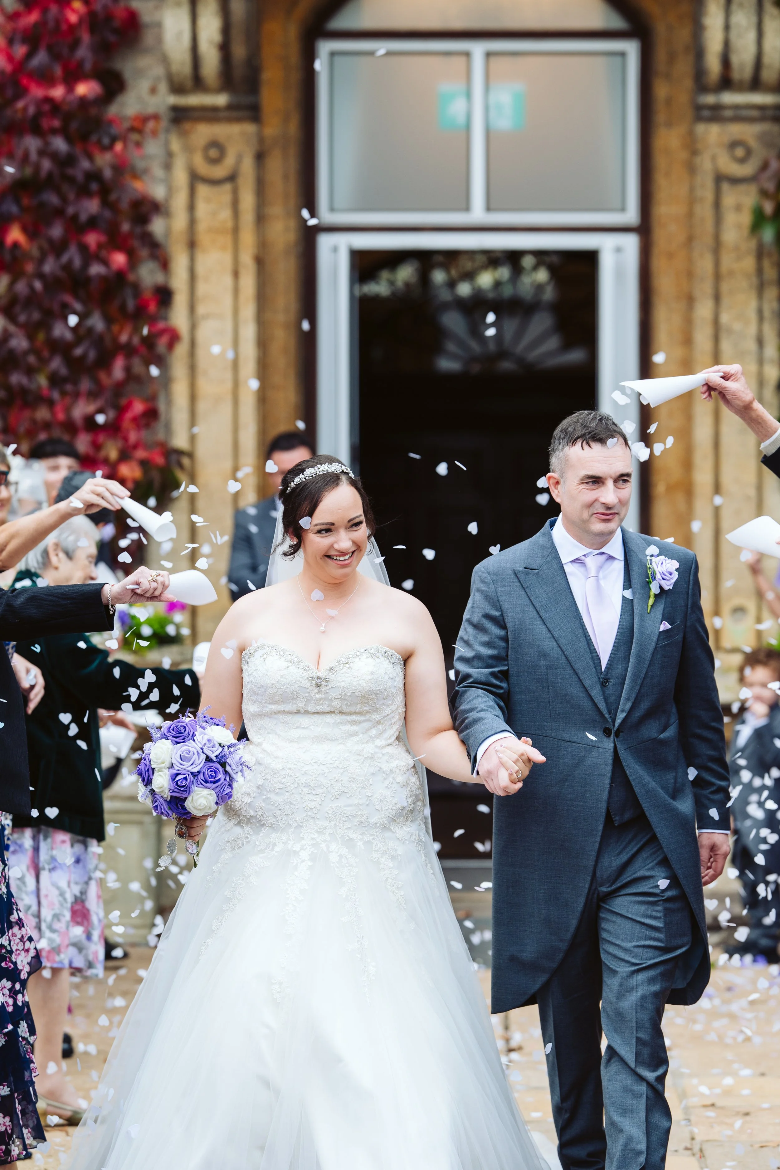 A bride and groom walking out of a wedding venue, surrounded by guests throwing confetti. The bride is holding a purple and white bouquet, wearing a strapless white wedding dress. The groom is dressed in a gray suit with a purple boutonniere. They are smiling and holding hands as confetti falls around them.