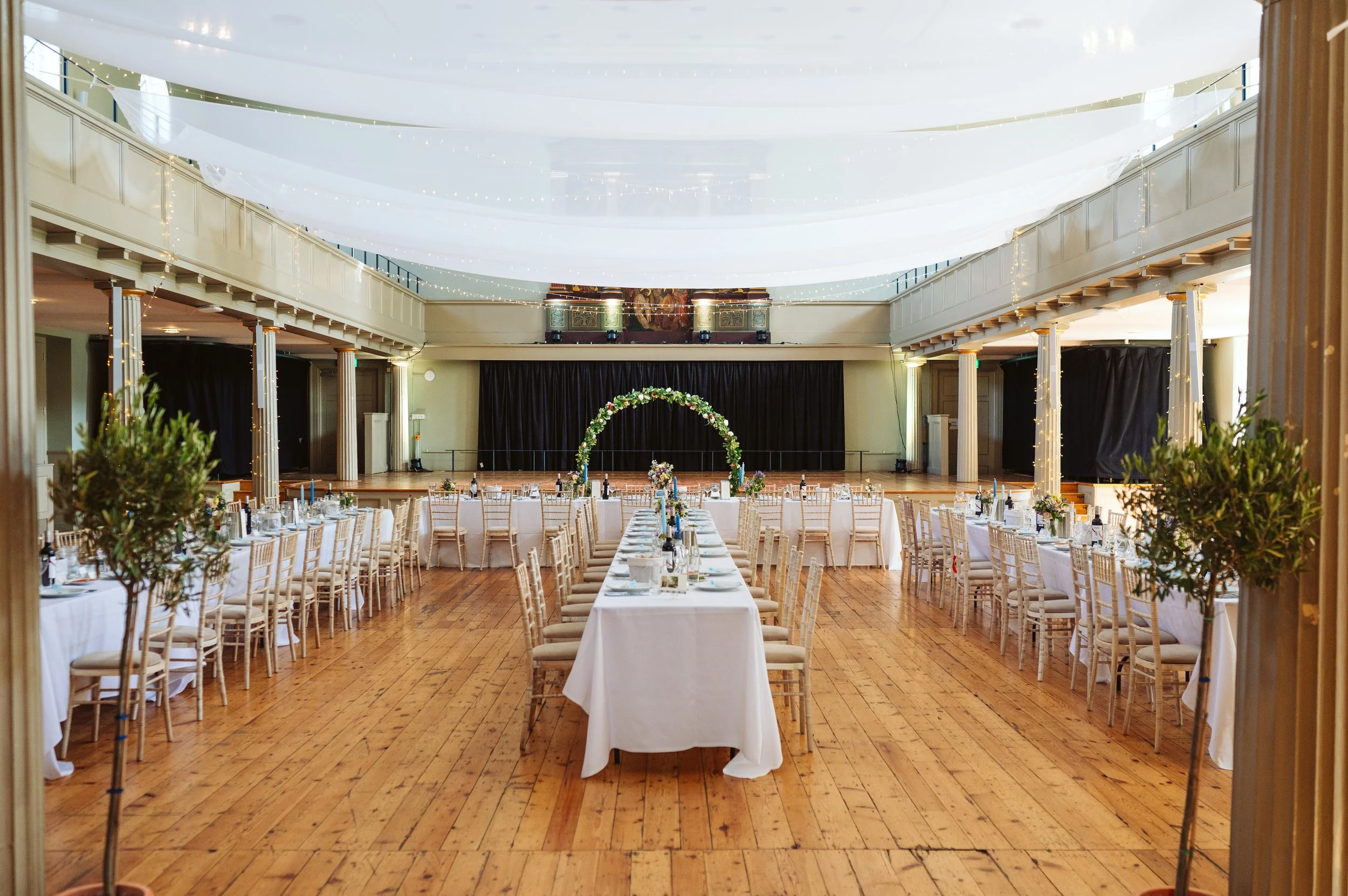 Elegantly decorated indoor event hall with long dining tables set with white tablecloths, chairs, and floral centerpieces. At the front, there is a floral arch on a stage with a black curtain backdrop, and string lights are hanging from the ceiling.