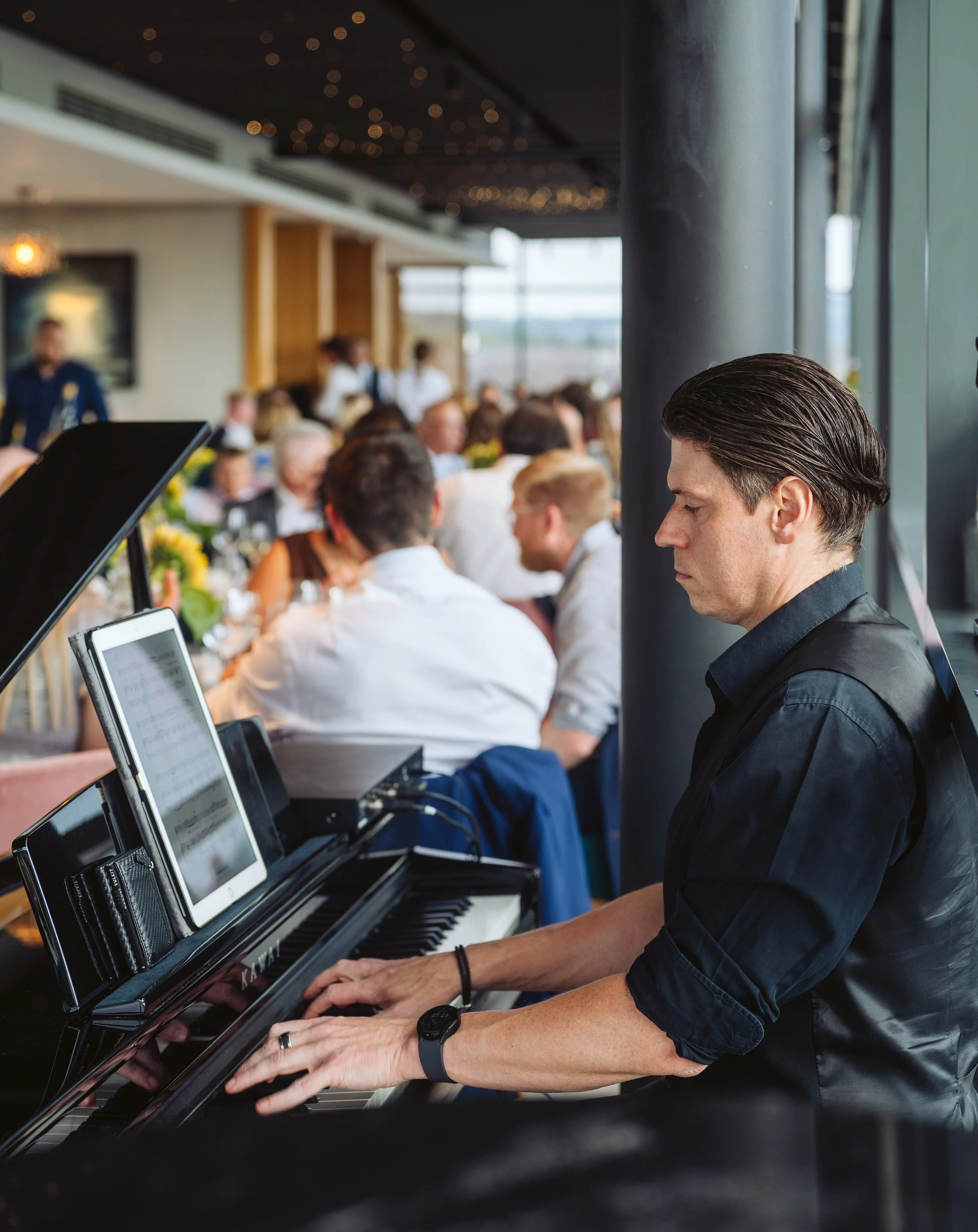 A man playing a black grand piano at a formal event with a crowd of people seated at tables in the background.