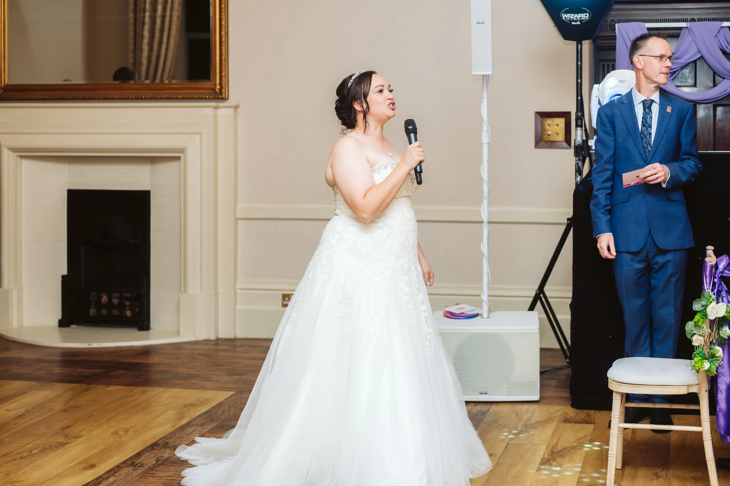 Bride in a white wedding dress holding a microphone, standing in a decorated reception hall.