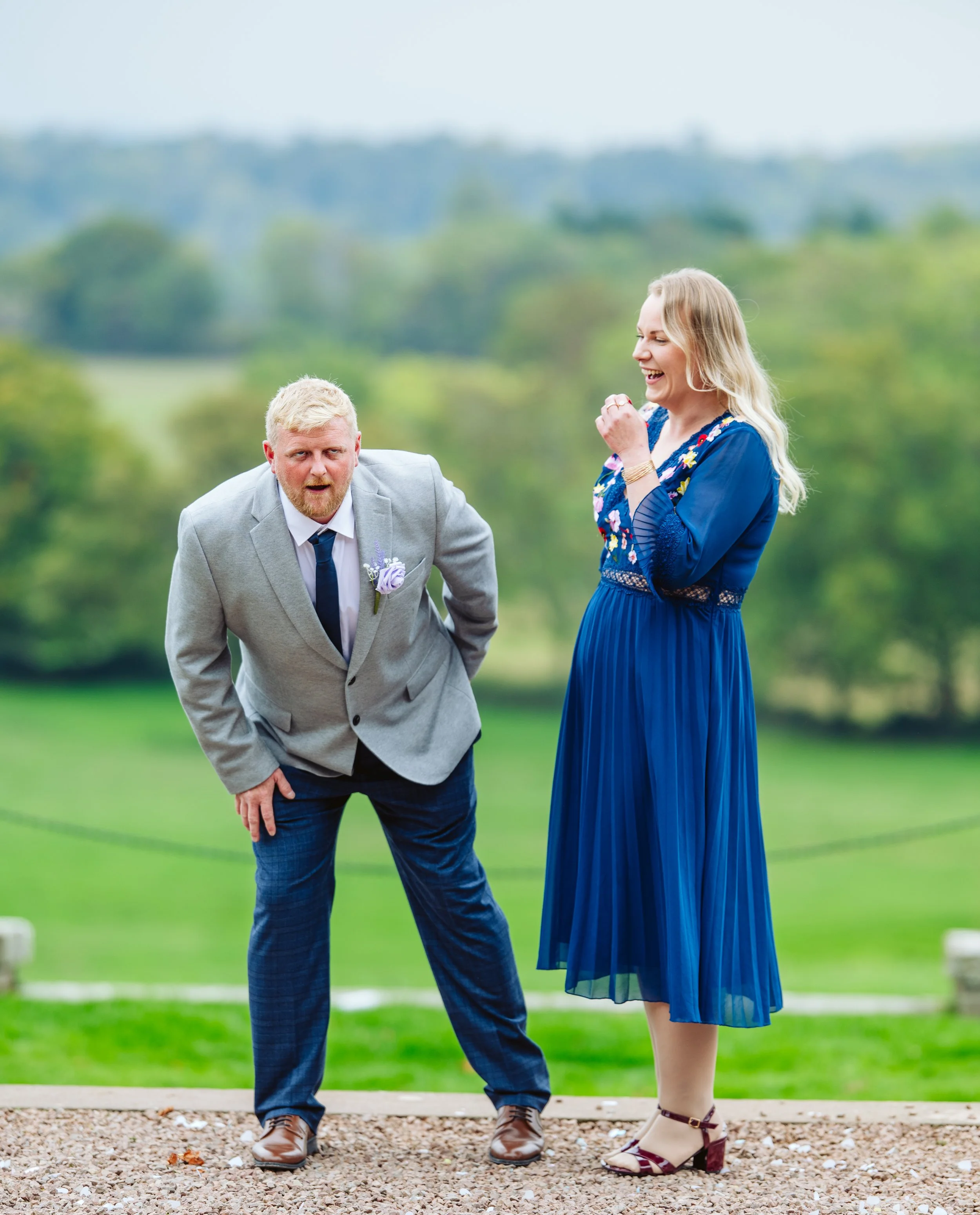 A man in a gray suit and a woman in a blue dress laughing outdoors on a grassy field with trees in the background.