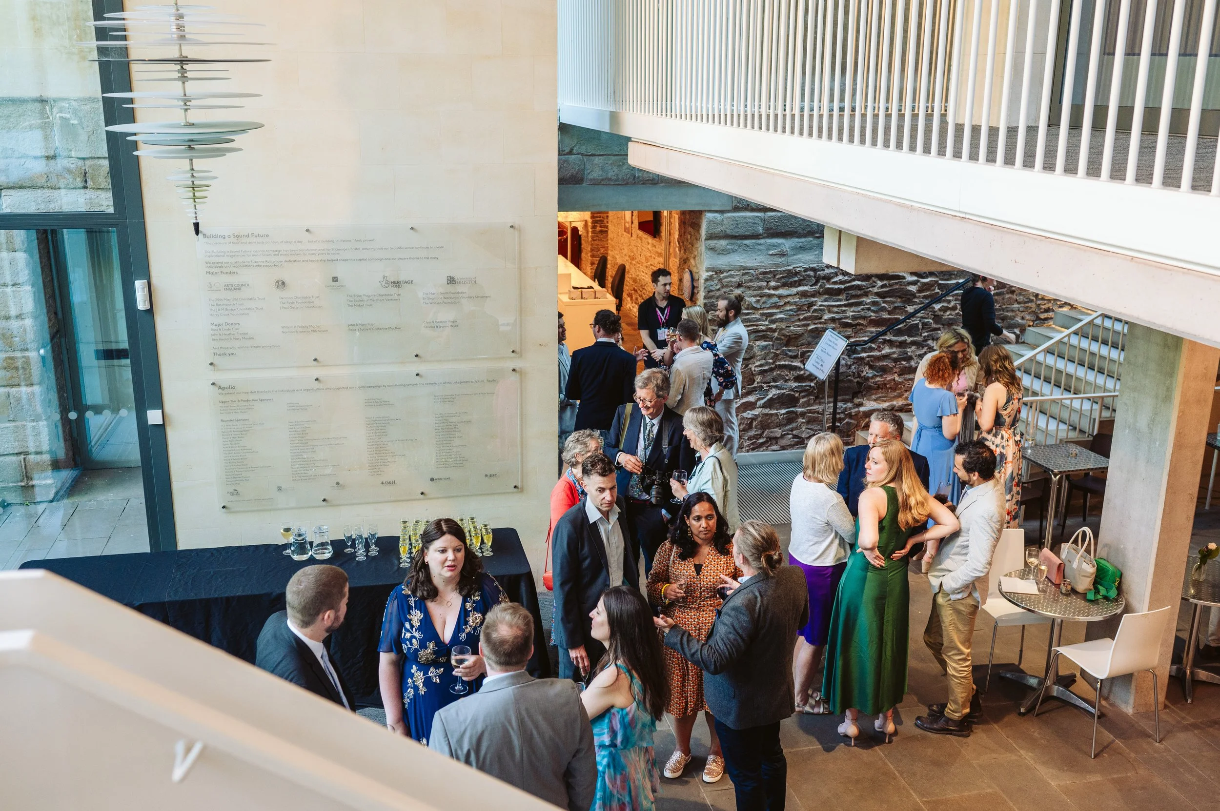 People gathered at a social event or reception inside a modern building with stone and glass walls, engaged in conversations. Some are holding drinks, and there are tables with bottles and glasses. The setting has a contemporary design with stairs and metal railings.