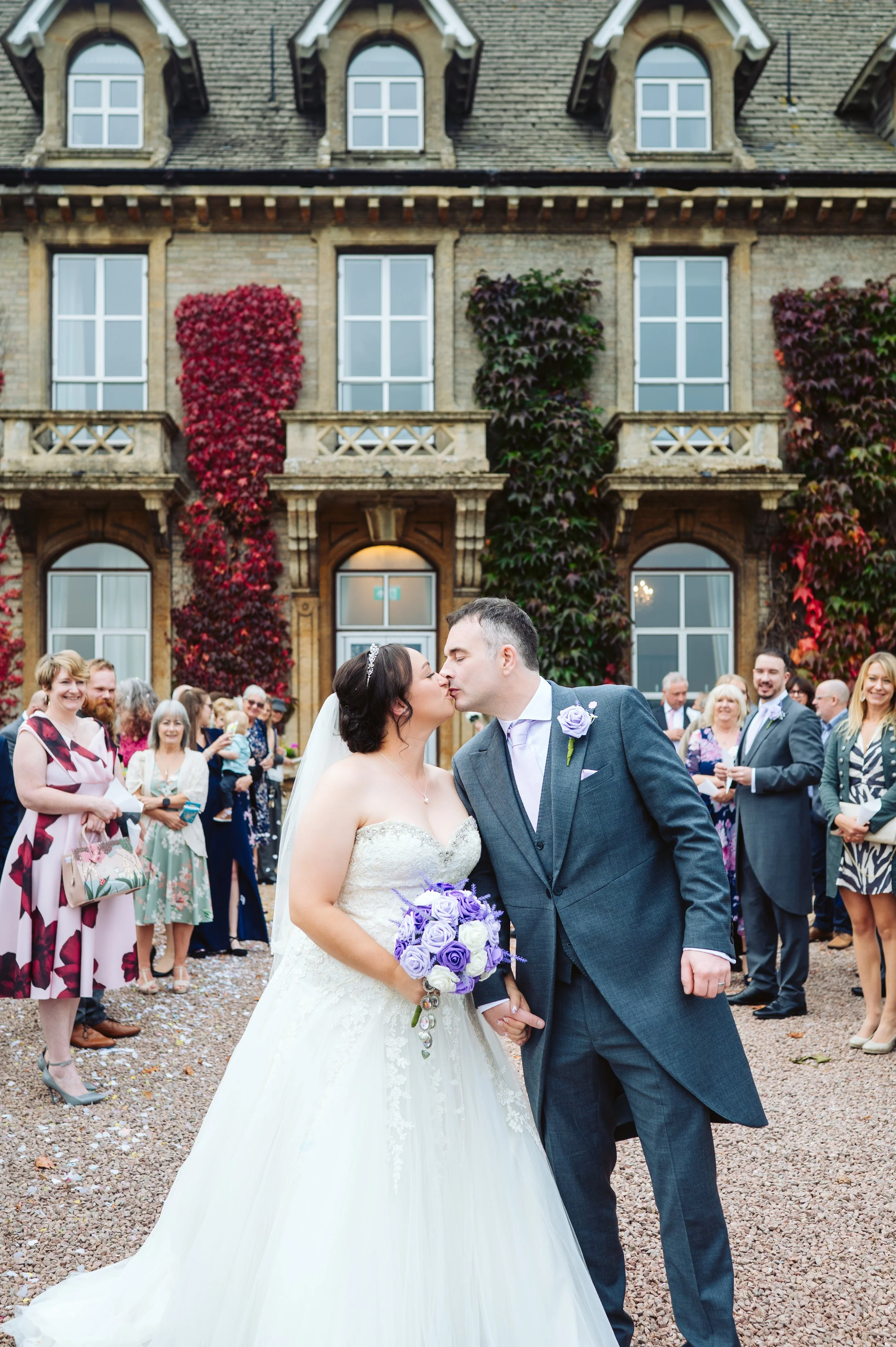 Bride and groom sharing a kiss in front of wedding guests, outside an ivy-covered brick building.