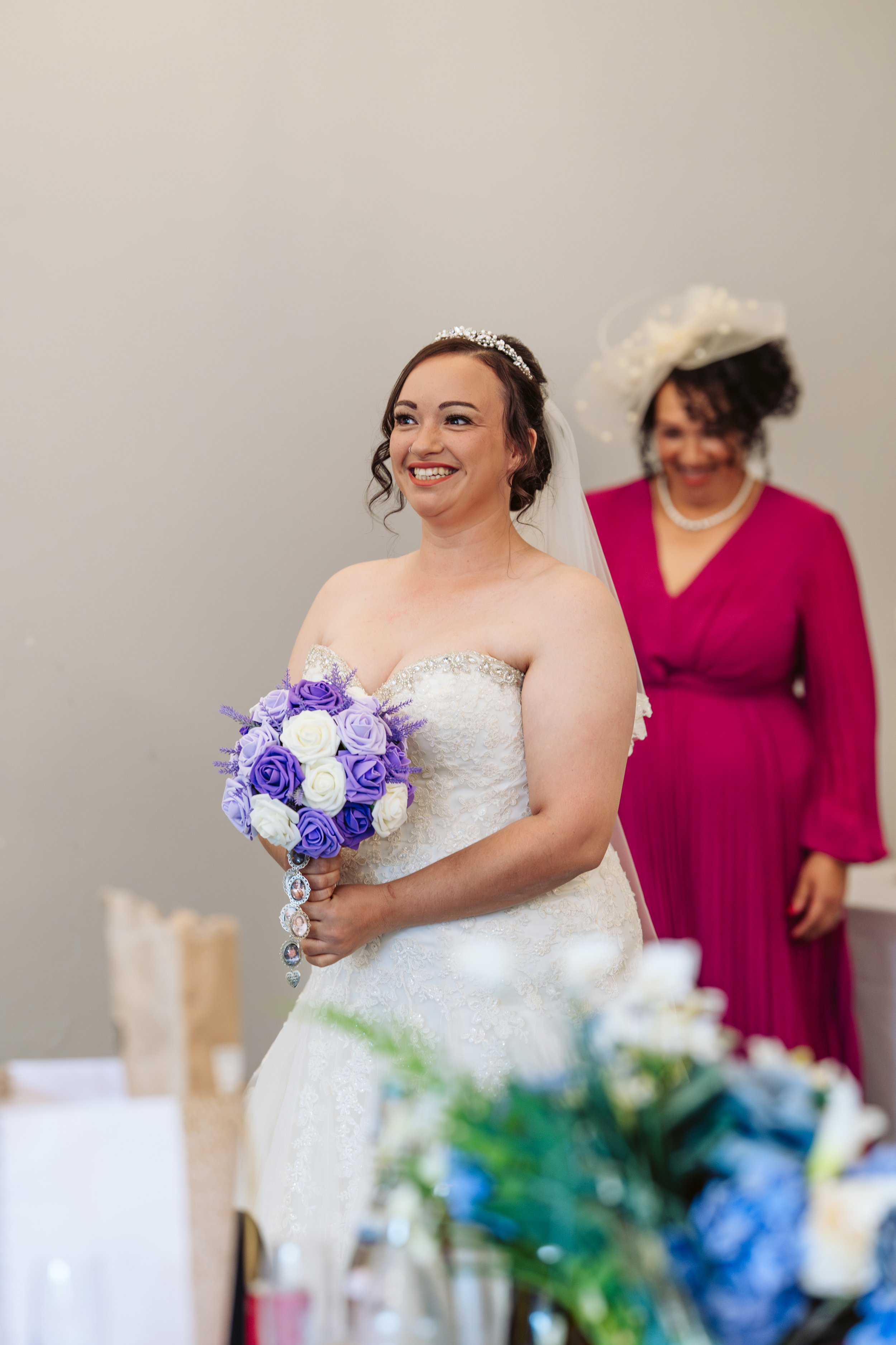 A bride in a strapless white wedding gown holding a bouquet of purple and white roses, smiling. An older woman in a magenta dress and large hat stands in the background, also smiling.