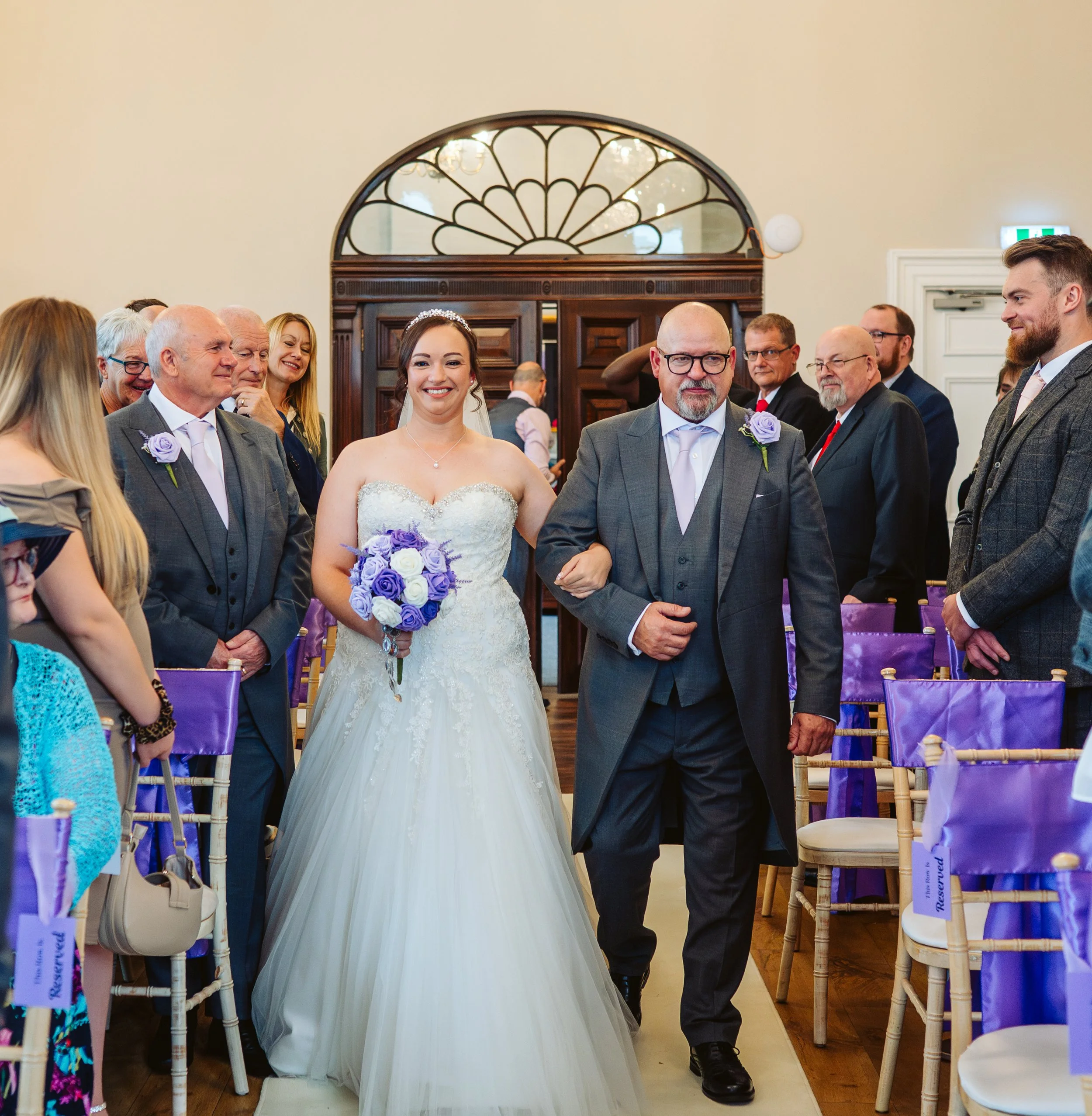 Bride walking down the aisle with a man, surrounded by seated guests at a wedding ceremony.