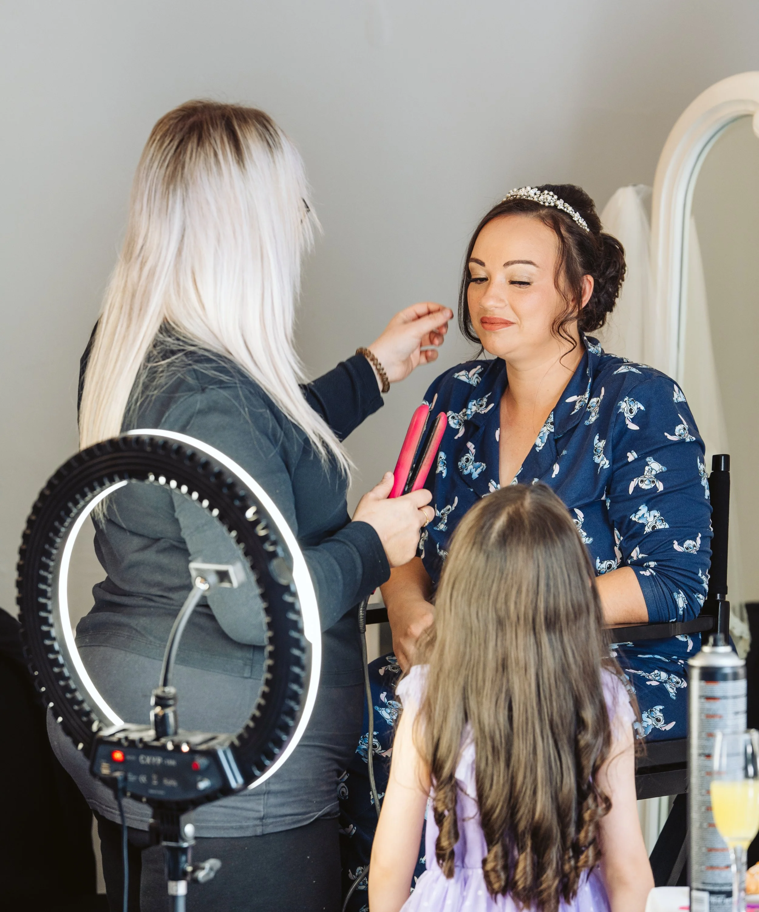 A woman in a blue floral robe is getting her makeup done by a makeup artist, with a little girl watching. The makeup artist holds styling tools, and there is a ring light in the foreground.
