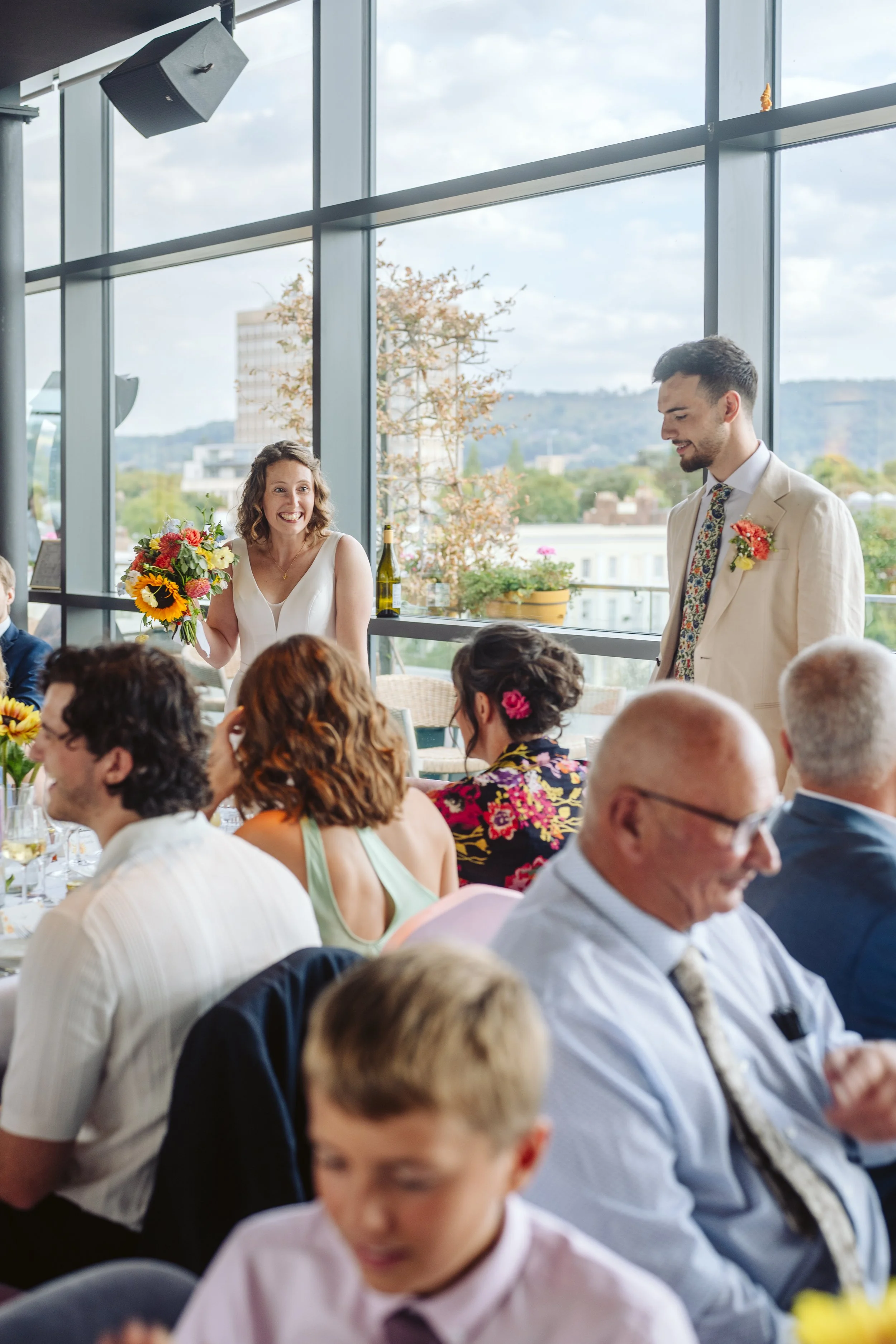 A woman holding a bouquet of colorful flowers stands and smiles as a man in a beige suit and floral tie stands nearby during a celebration, with others seated at tables in a bright room with floor-to-ceiling windows and an outdoor view.