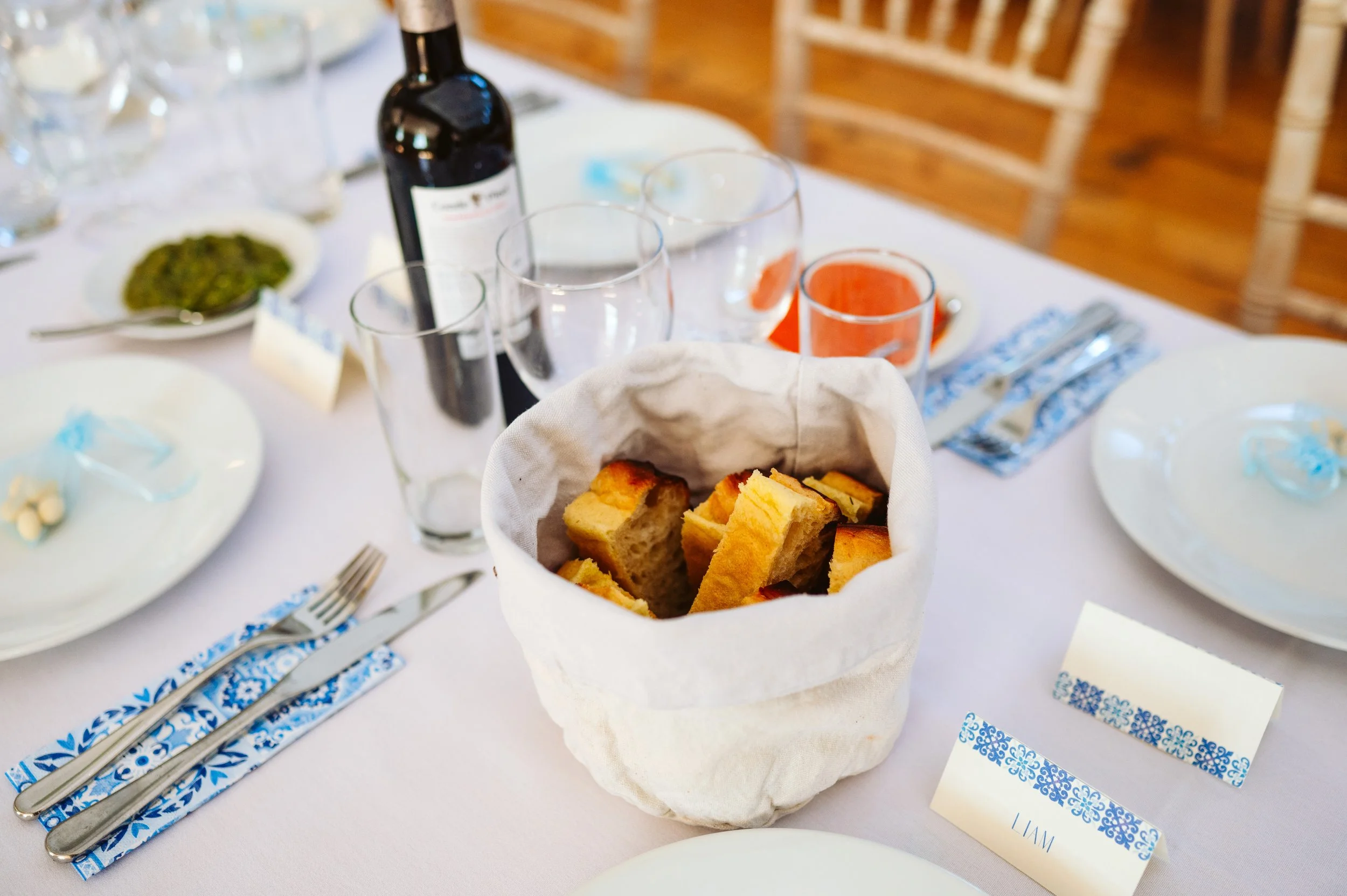 A table set for a meal with a basket of bread, wine, multiple glasses, plates, and utensils.