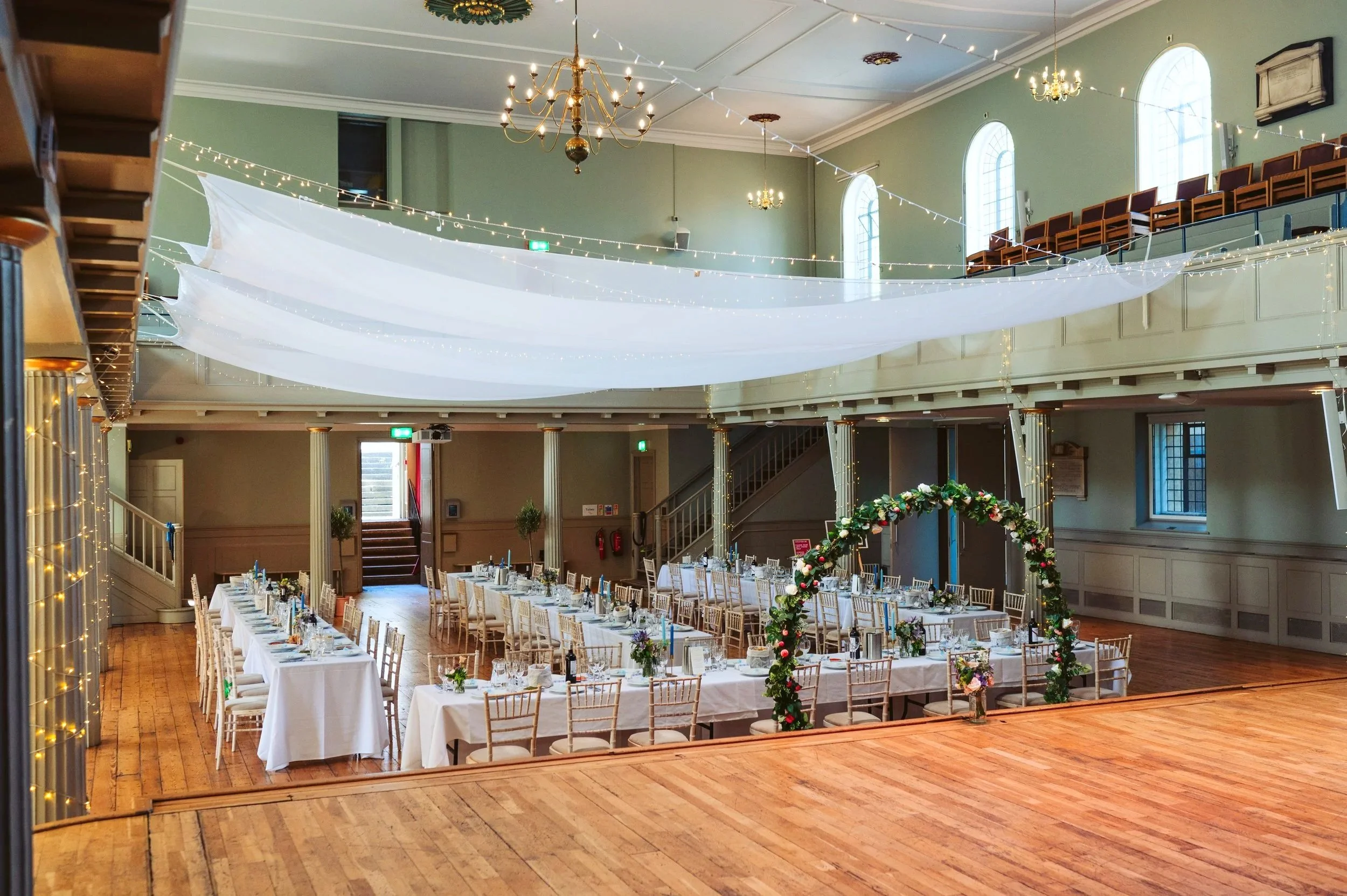 Event hall decorated for a wedding reception with long tables, floral arrangements, and an archway, with draped fabric and string lights hanging from the ceiling.