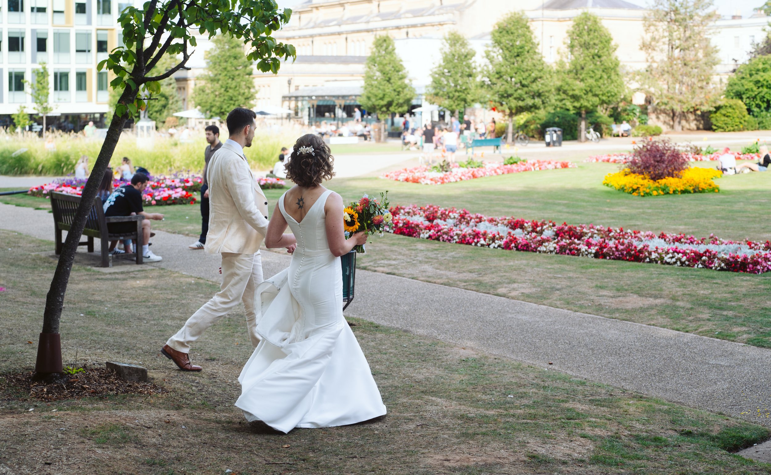 A bride and groom walking in a park with blooming flowers and trees, surrounded by people sitting on benches and enjoying the day.