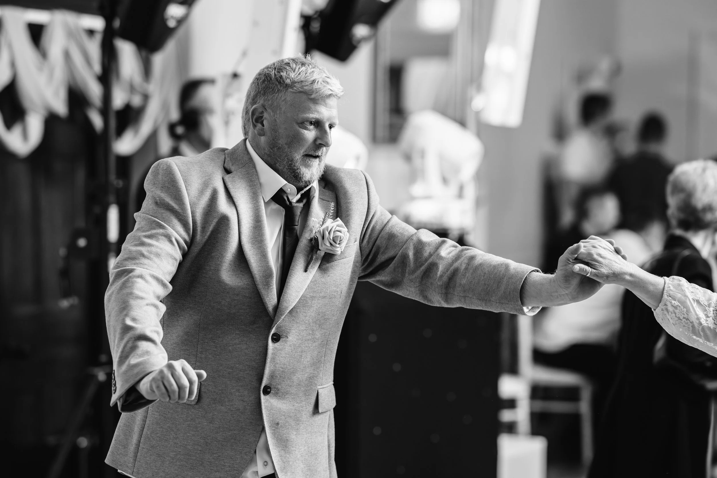 A man with a beard in a suit and tie, with a flower pinned to his jacket, dancing with a woman at a wedding reception in a black-and-white photograph.