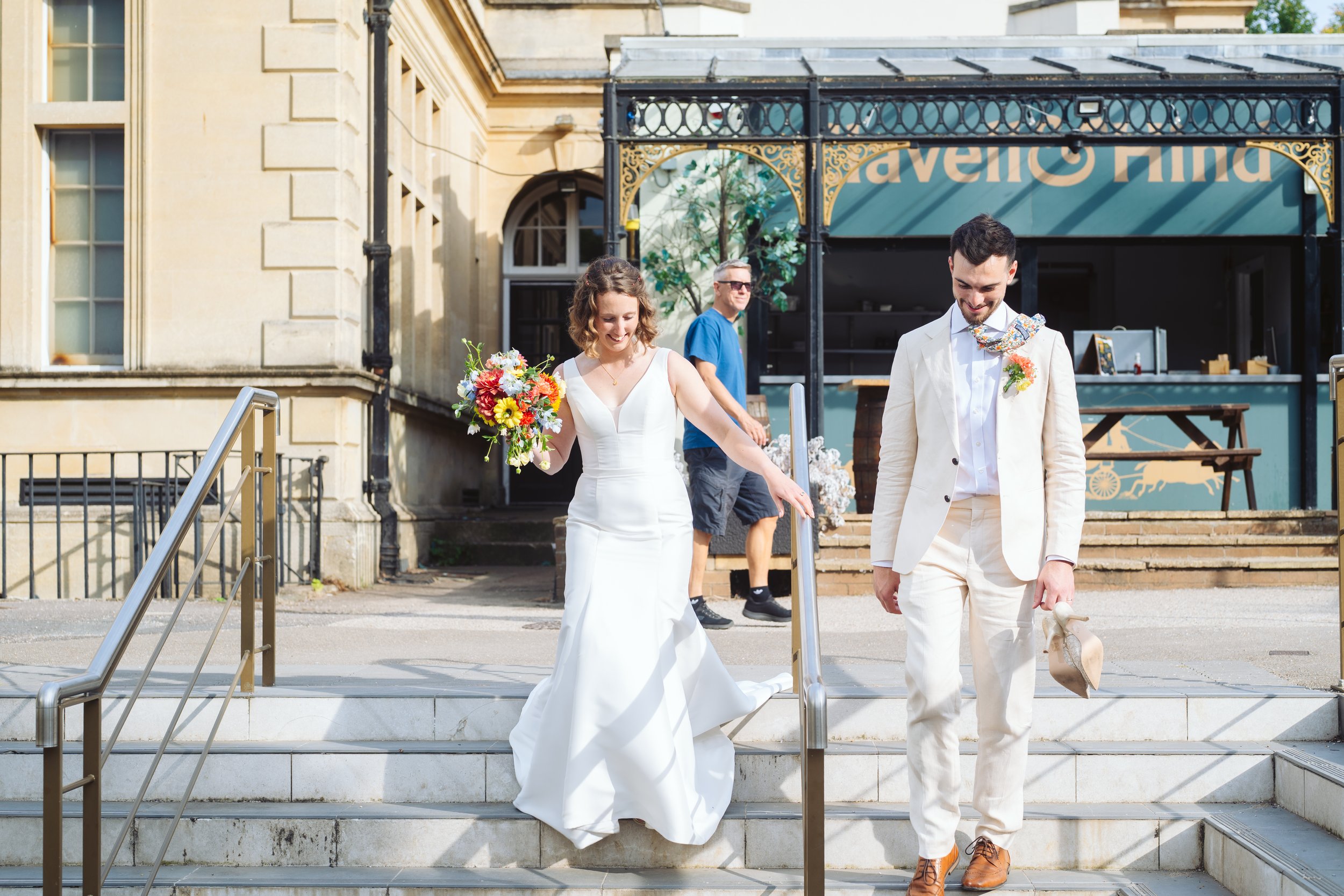 A bride and groom walking down the steps outside a building, with a person in a blue shirt in the background