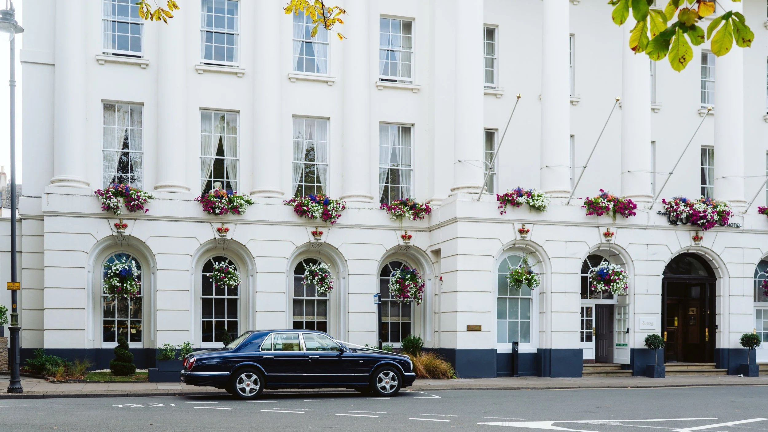 White hotel building with arched windows and colorful flower arrangements, parked black luxury car in front.