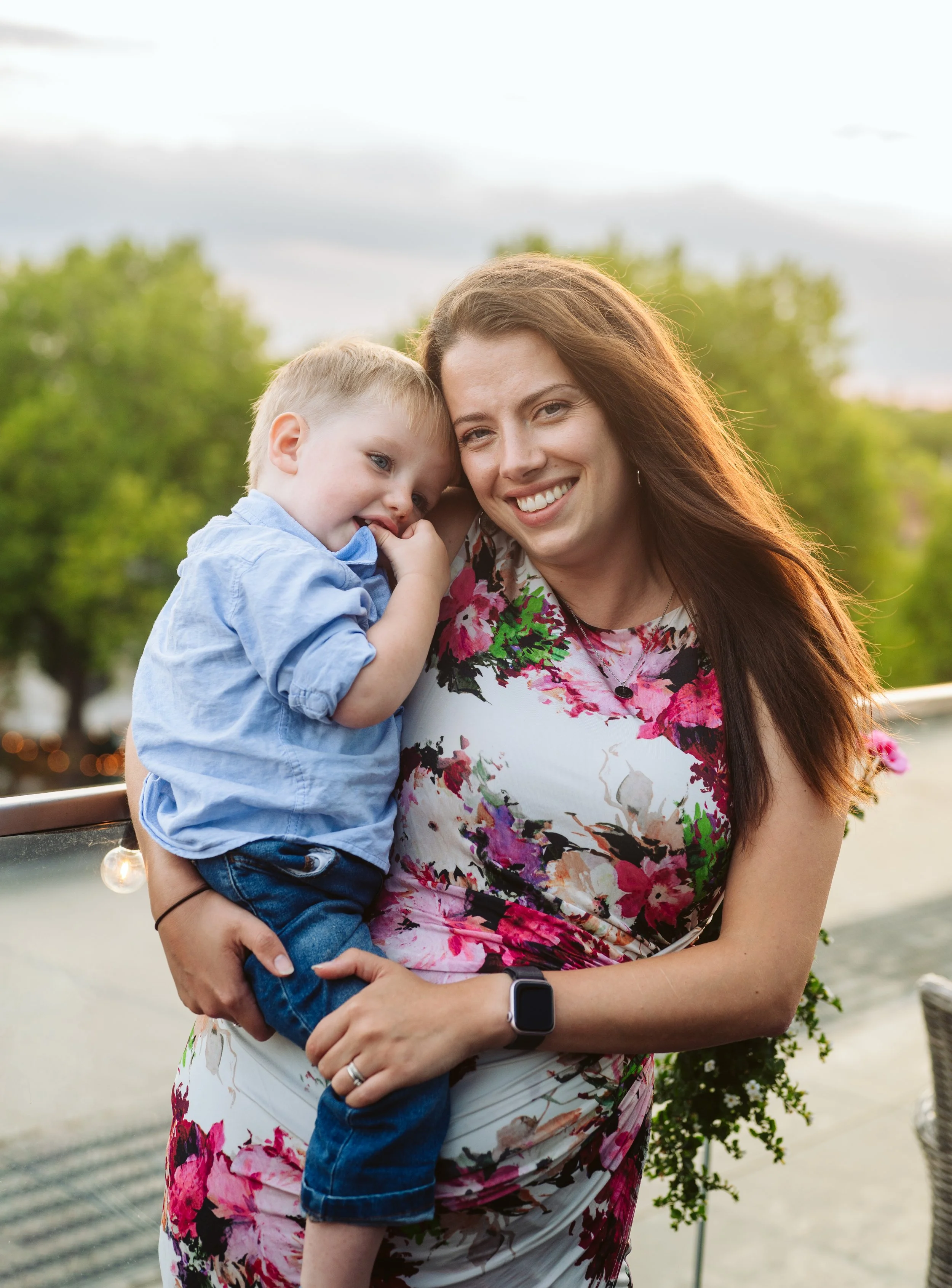 A woman holding a young boy, both smiling, outdoors with green trees and a cloudy sky in the background.