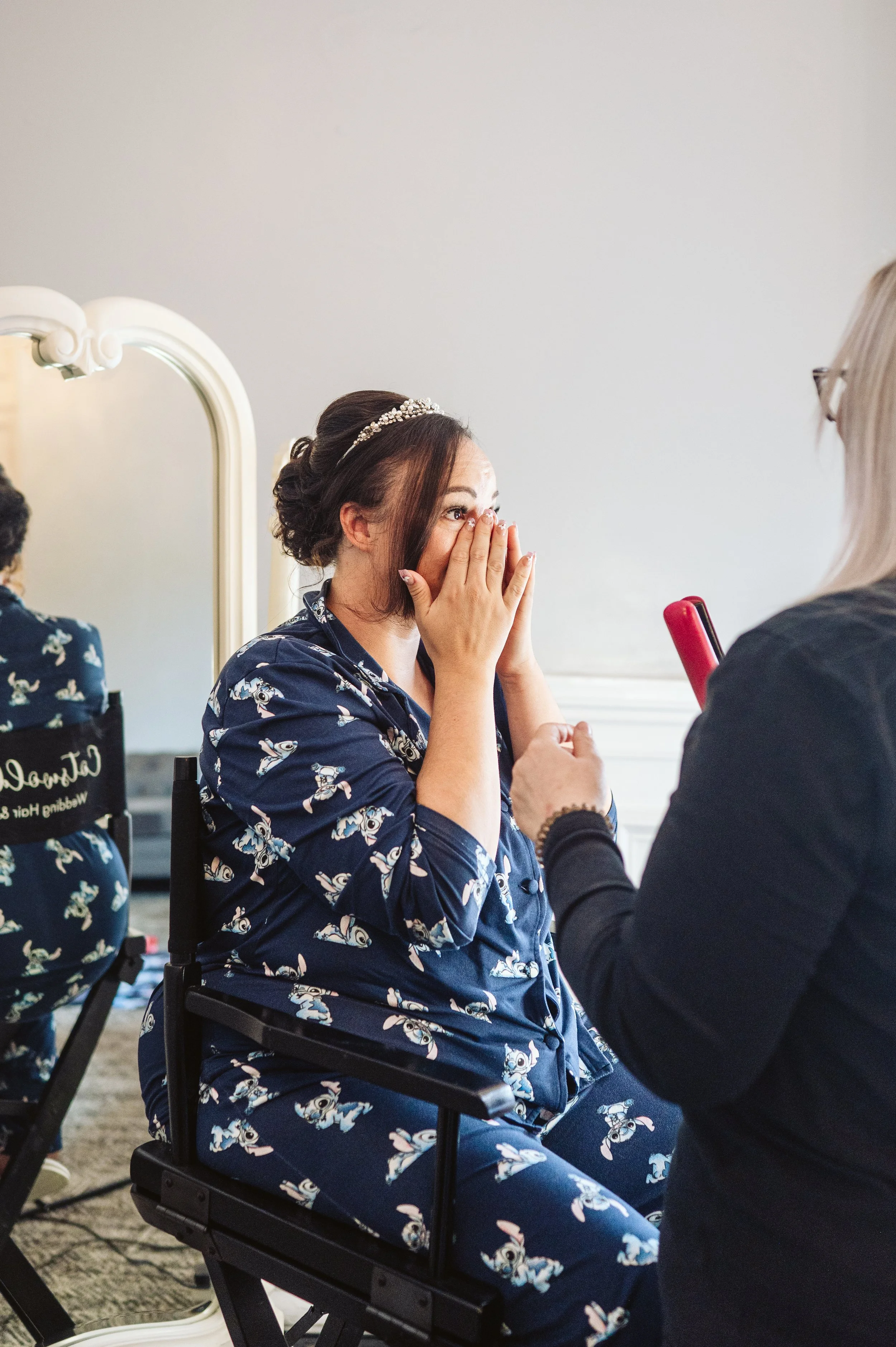 A woman sitting in a chair looking surprised with her hands covering her mouth while a stylist or makeup artist works on her face, in a room with a mirror.