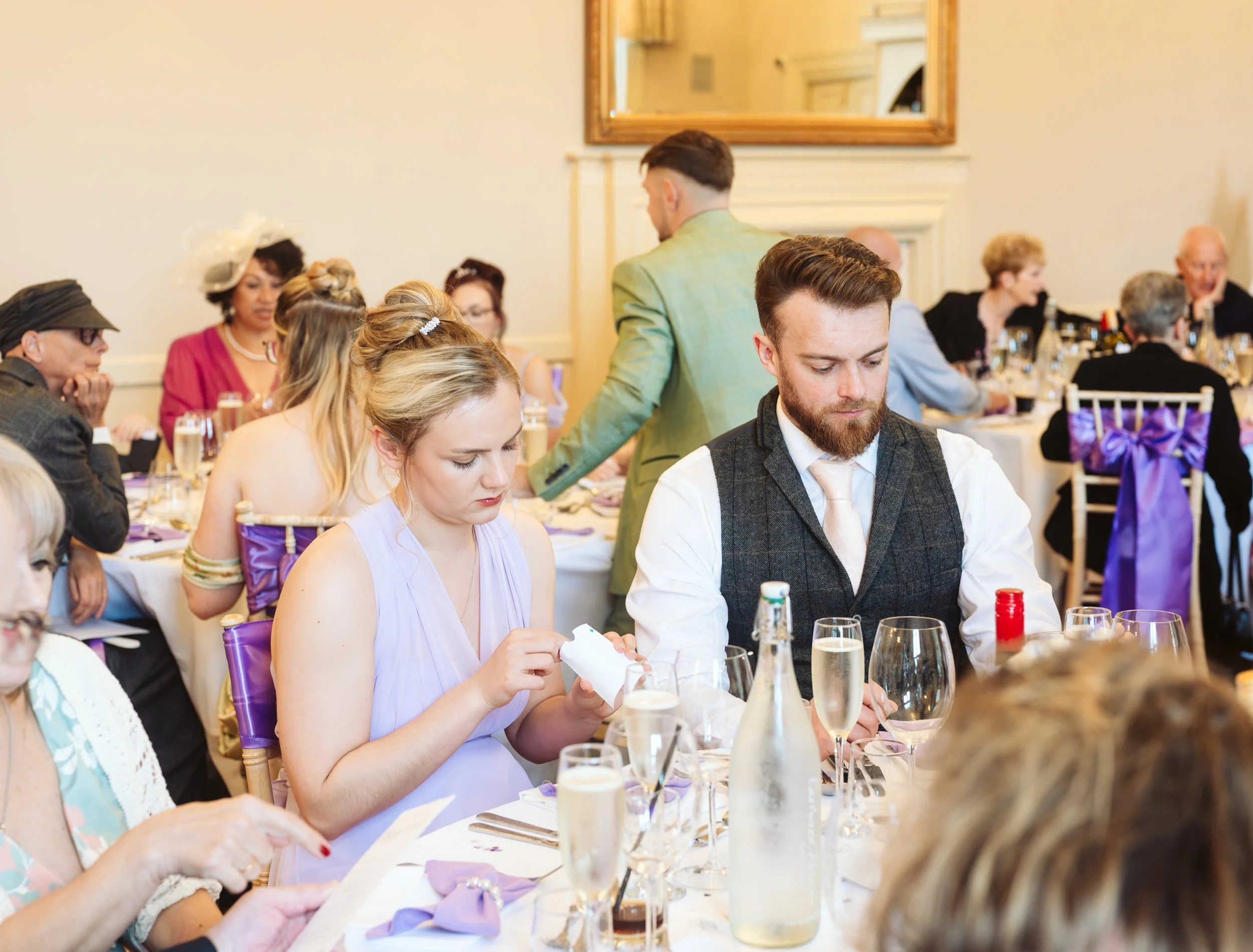 People sitting at banquet tables during a formal event, with some looking at programs and others writing, decorated with purple ribbons on chairs, in a well-lit room with a fireplace and large mirror.