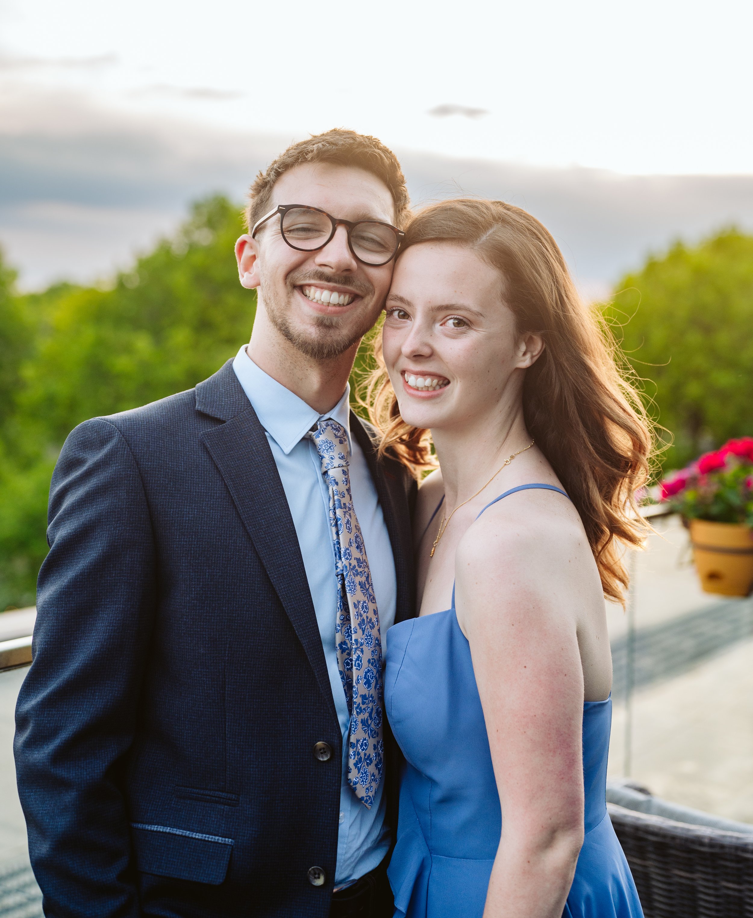 A smiling young man and woman standing close together outdoors during sunset. The man is wearing glasses, a navy suit, and a patterned tie. The woman has long, wavy hair and is wearing a blue dress. They are embracing and appear happy.