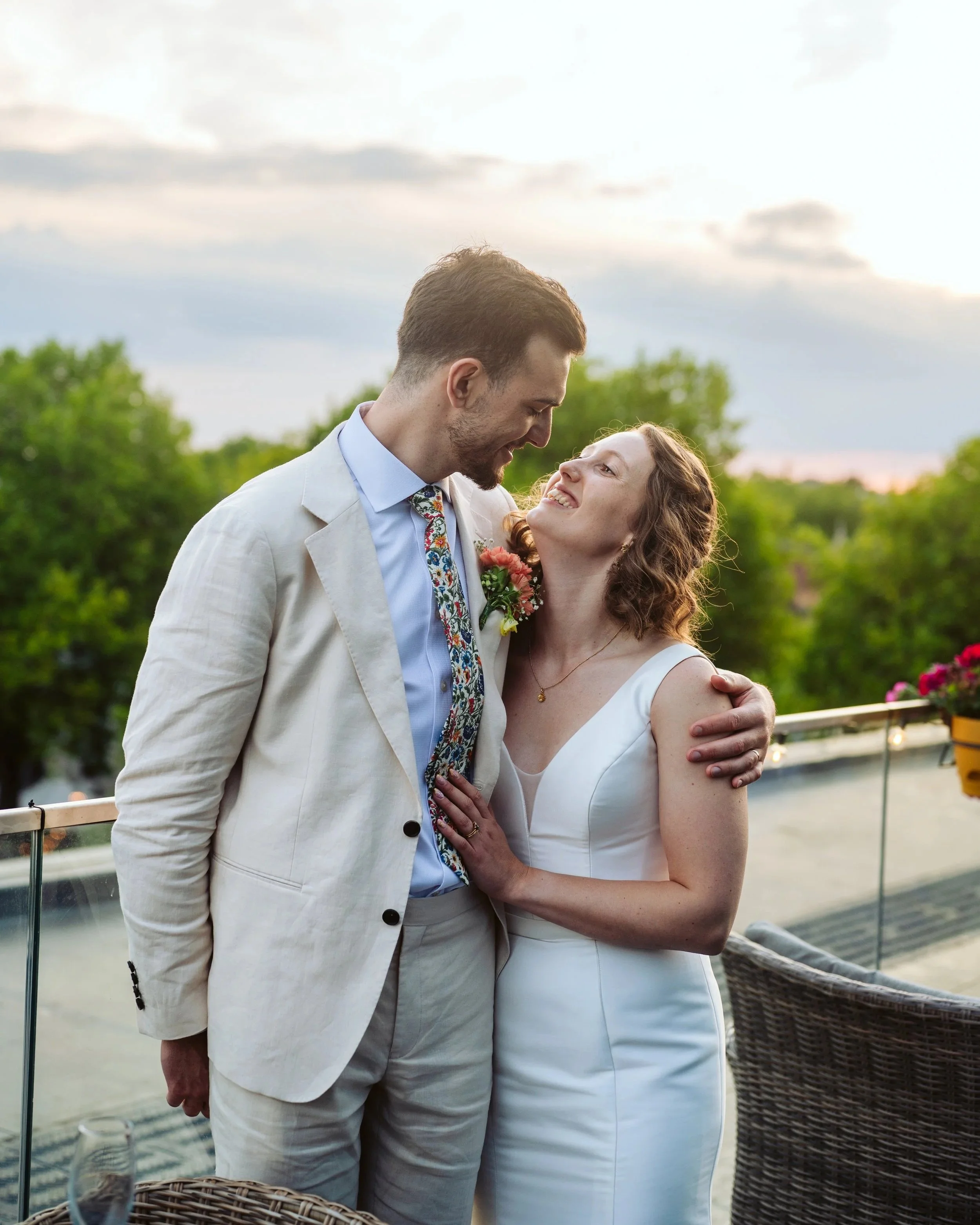 Couple in wedding attire on a balcony, smiling and looking into each other's eyes, with greenery and a cloudy sky in the background.