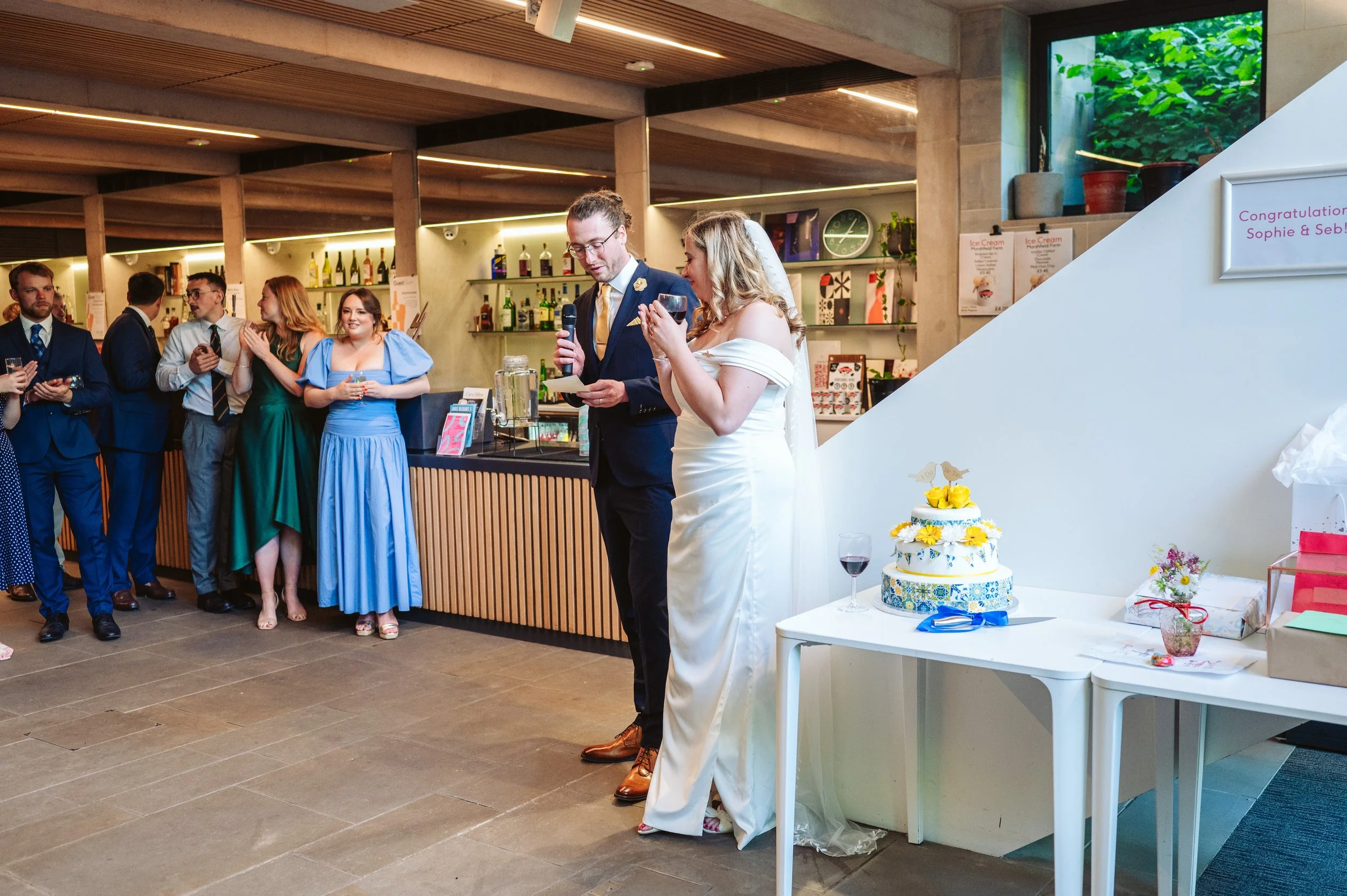 Bride and groom giving a speech at their wedding reception, with guests listening. The bride is in a white off-shoulder gown holding a glass of wine, and the groom is in a dark suit speaking into a microphone. A decorated wedding cake with yellow flowers is on a small table nearby. In the background, guests are dressed in formal attire, some holding glasses, and a bar area is visible.