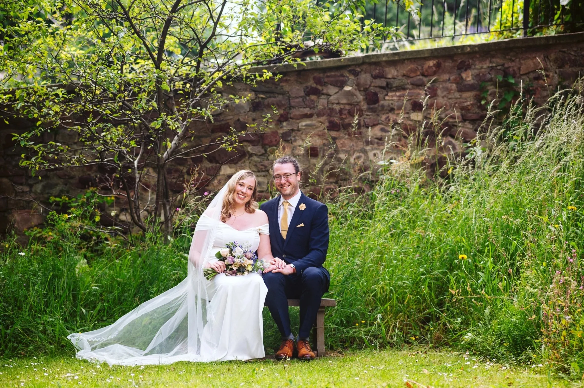 A happy bride and groom sitting on a small bench outdoors, surrounded by green grass, trees, and wildflowers, with a stone wall in the background.