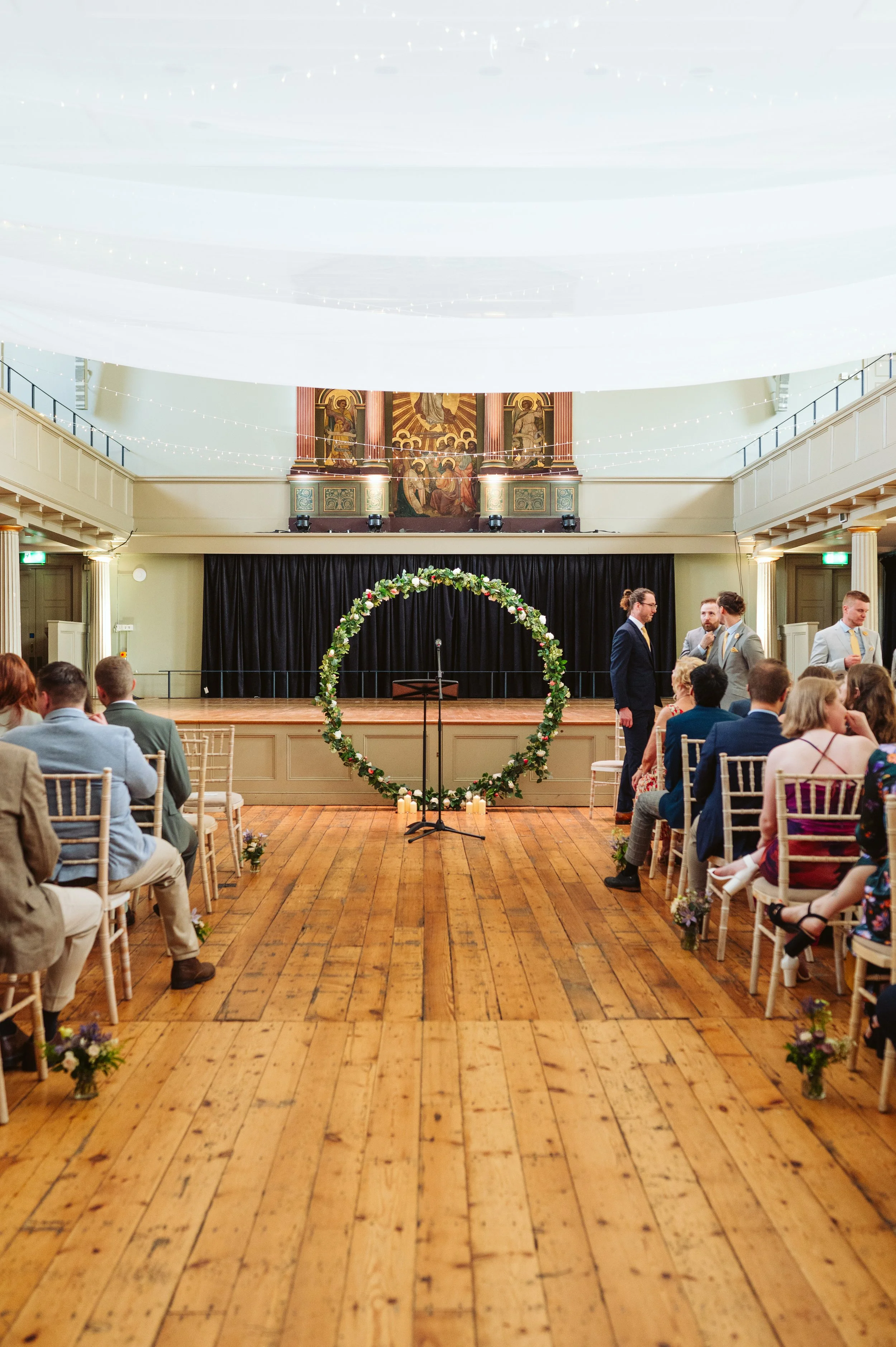 Indoor wedding ceremony setup with rows of chairs, a floral arch on a wooden floor, and guests seated. A black curtain backdrop is on stage, above which are religious paintings. Candles line the floor in front of the flower arch, and string lights are hanging from the ceiling.