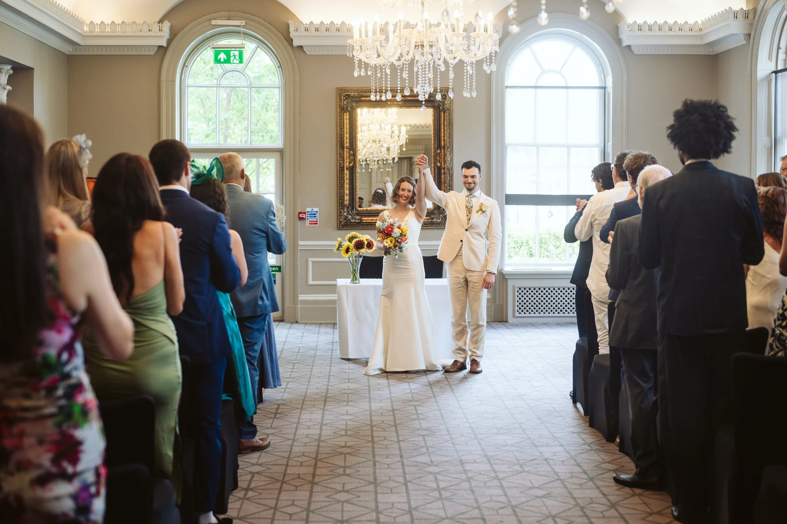 A bride and groom in wedding attire standing in a decorated banquet room, holding hands raised in celebration, with guests standing on either side, and large windows, chandelier, and mirror in the background.