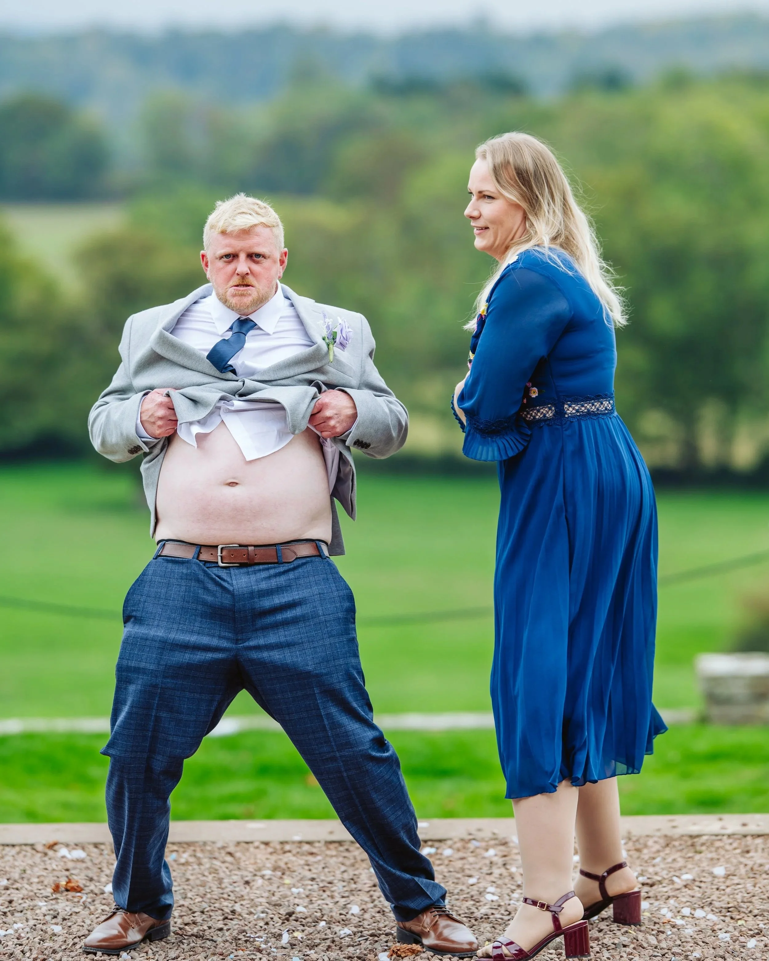 A man in a suit is lifting his shirt to reveal his stomach while standing outdoors, with a woman in a blue dress watching and smiling.