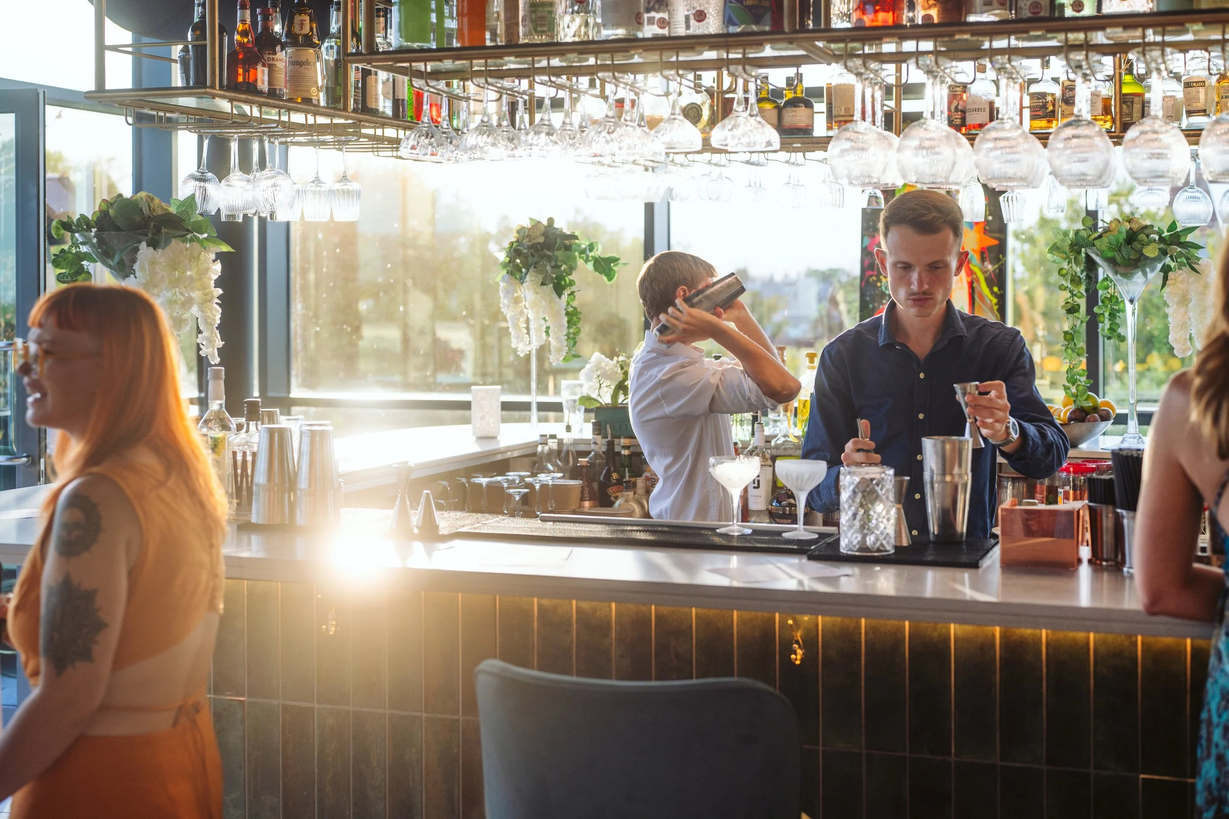 A bartender mixes drinks behind a bar as a man prepares a cocktail, with a woman with red hair and tattoos standing to the left, and a woman with blonde hair partially visible to the right, in a bright, modern bar with large windows and decorative flowers.