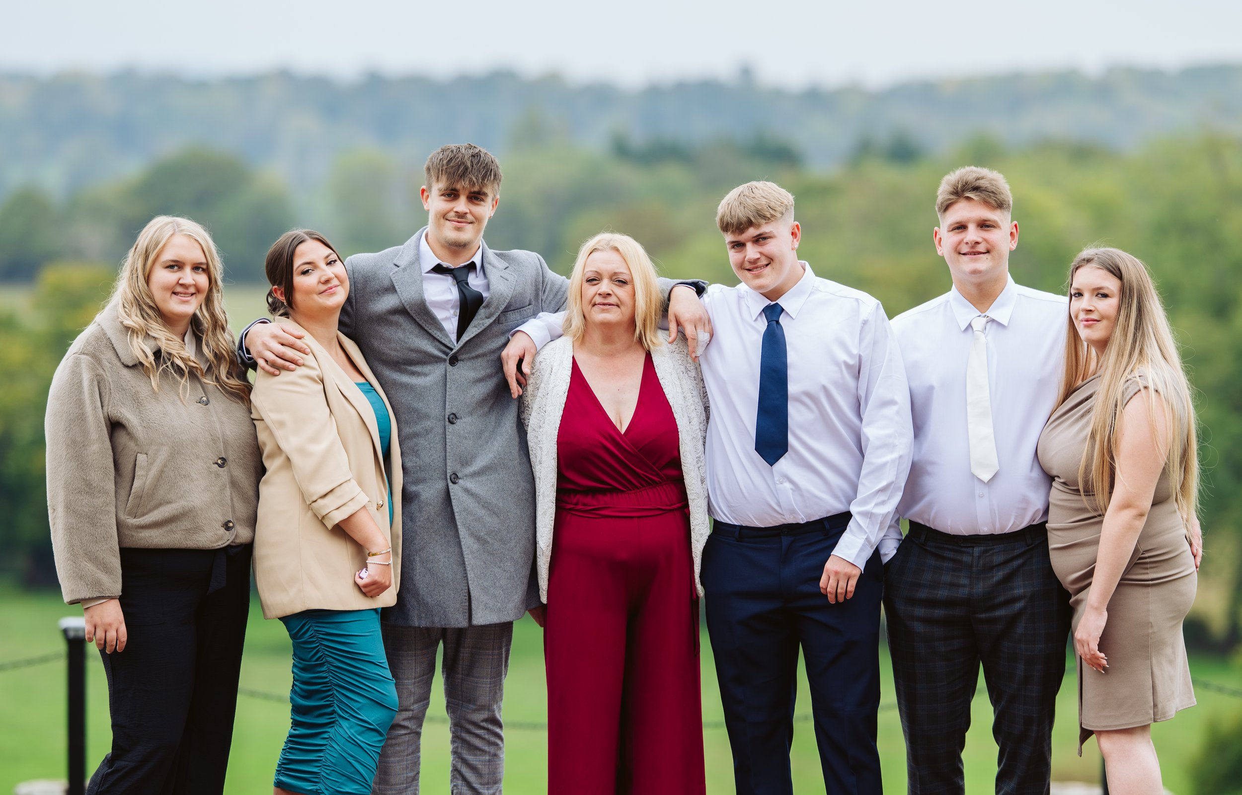 Family group of seven people, five women and two men, standing outdoors with green fields and trees in the background, smiling for the photo.