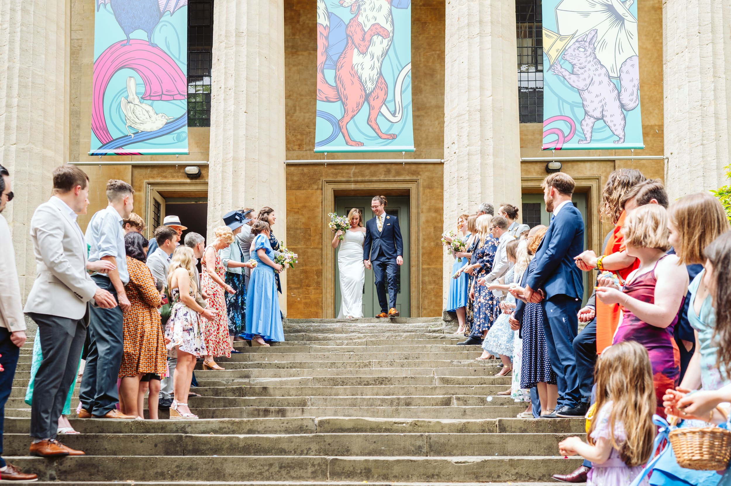 A bride and groom walking down the steps of a building, surrounded by friends and family, during a wedding celebration. The guests are standing in two lines, holding flowers or small items, and some are taking photos. The building has large columns and colorful banners with animal illustrations hanging above.