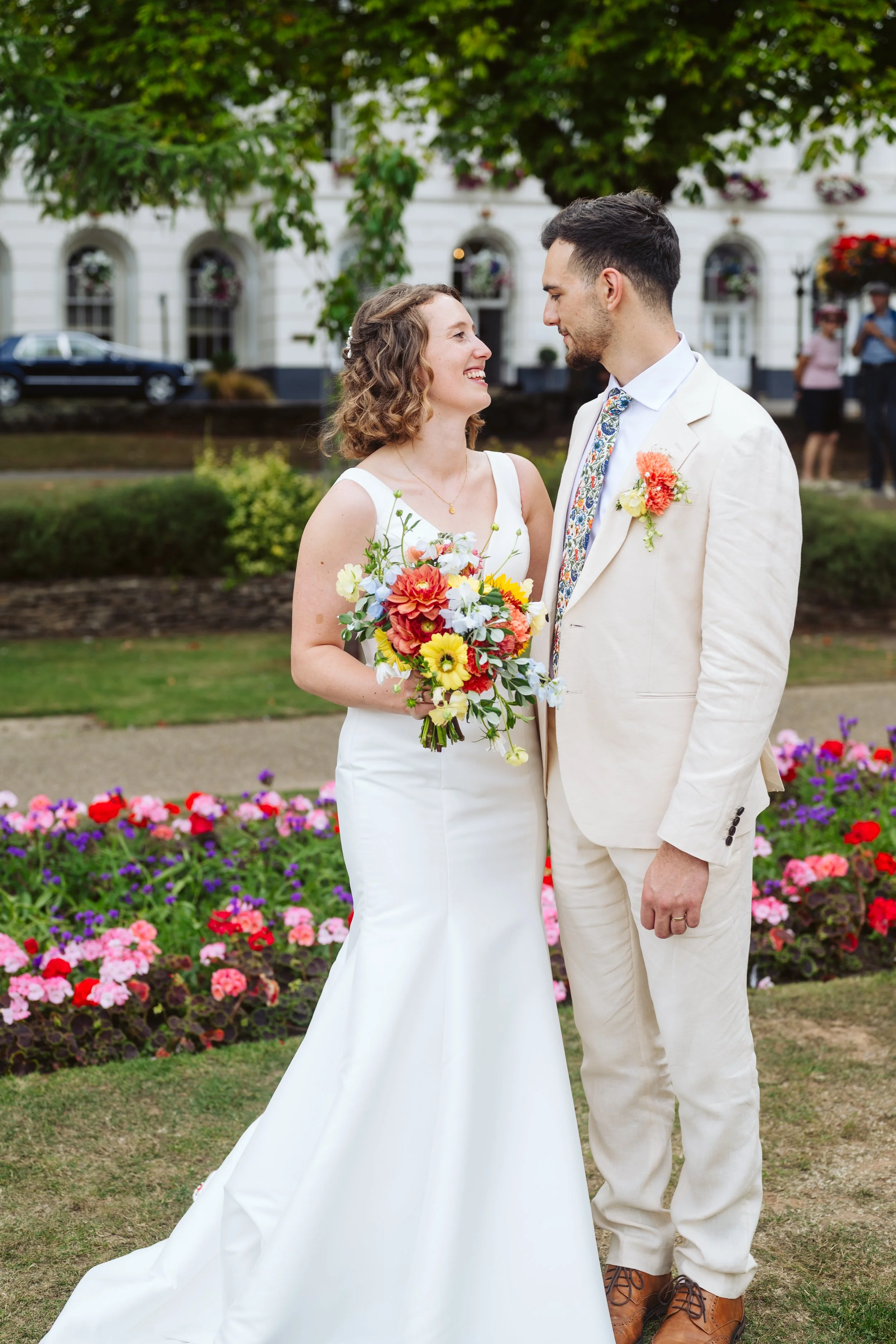 A bride and groom standing close, smiling at each other in a garden with colorful flowers. The bride is holding a bouquet of vibrant flowers and wearing a white wedding dress, while the groom is dressed in a light-colored suit with a floral boutonniere.