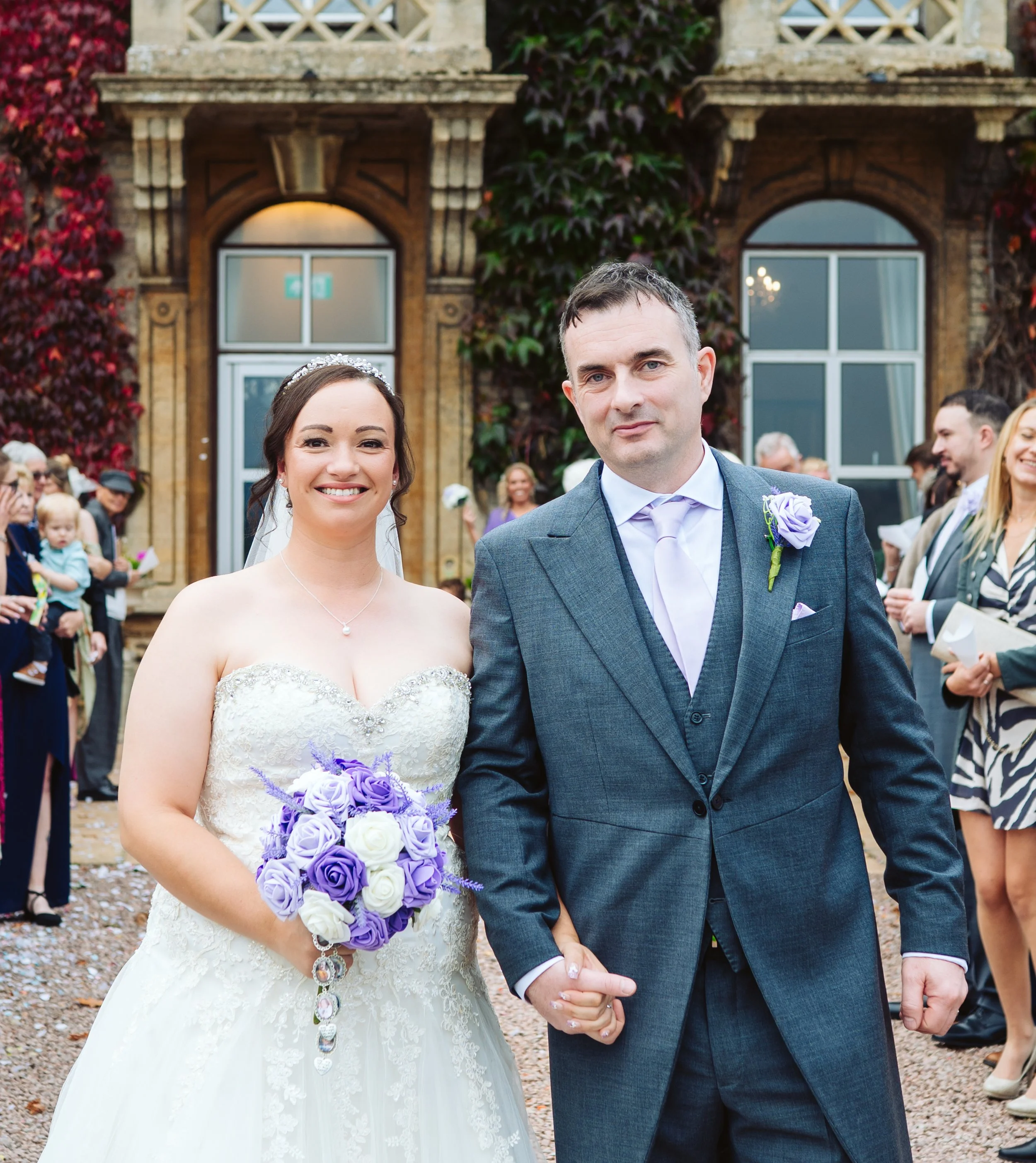 Bride and groom holding hands outdoors at their wedding, surrounded by guests, with a building covered in vines in the background.