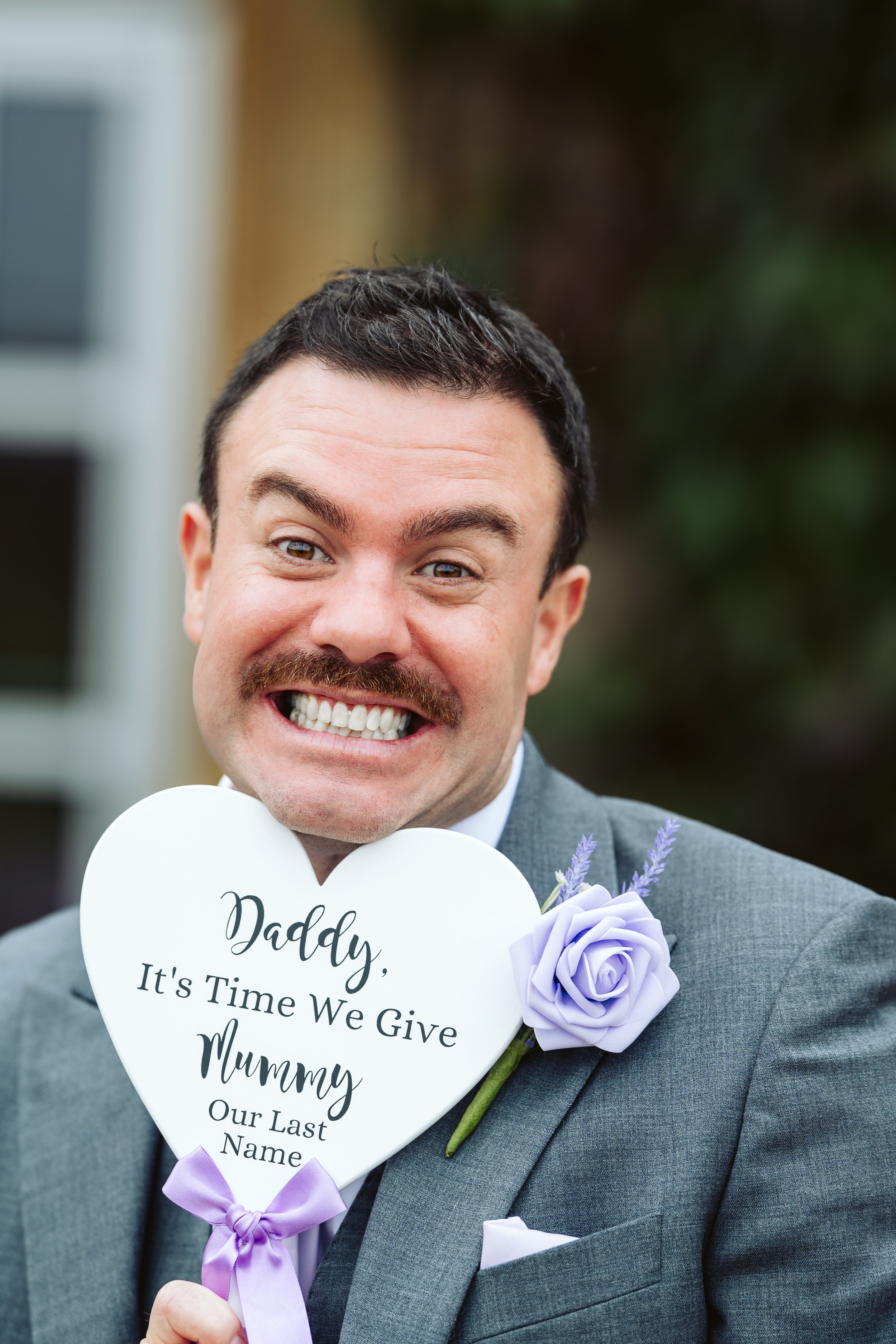 A man in a gray suit holding a white heart-shaped sign that reads 'Daddy, It's Time We Give Mummy Our Last Name,' with a purple ribbon and lavender flowers attached, smiling broadly.