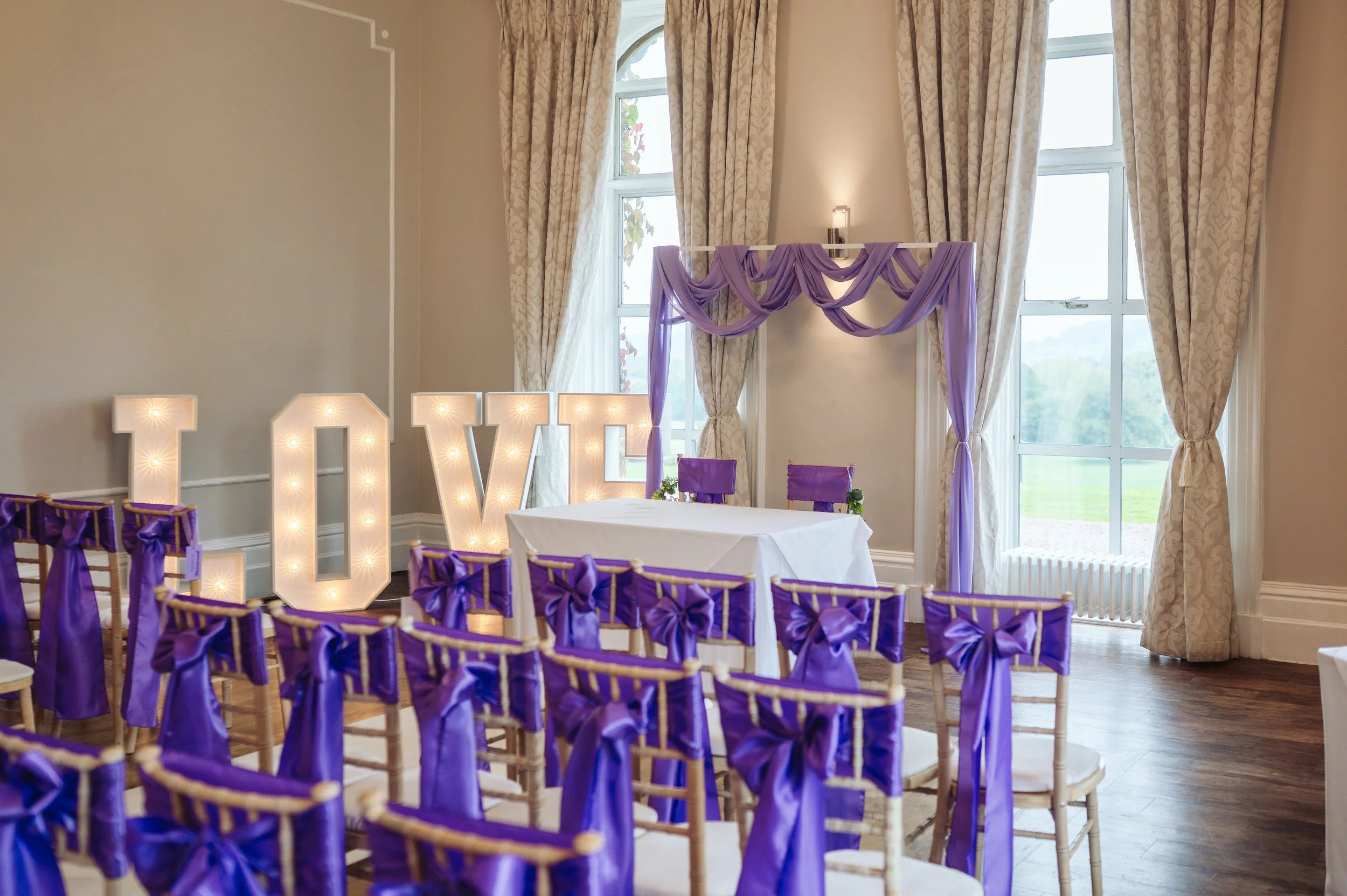 Wedding or event venue decorated with purple bows on chairs, purple drapes, and large illuminated letters spelling 'LOVE' near windows with cream-colored curtains.