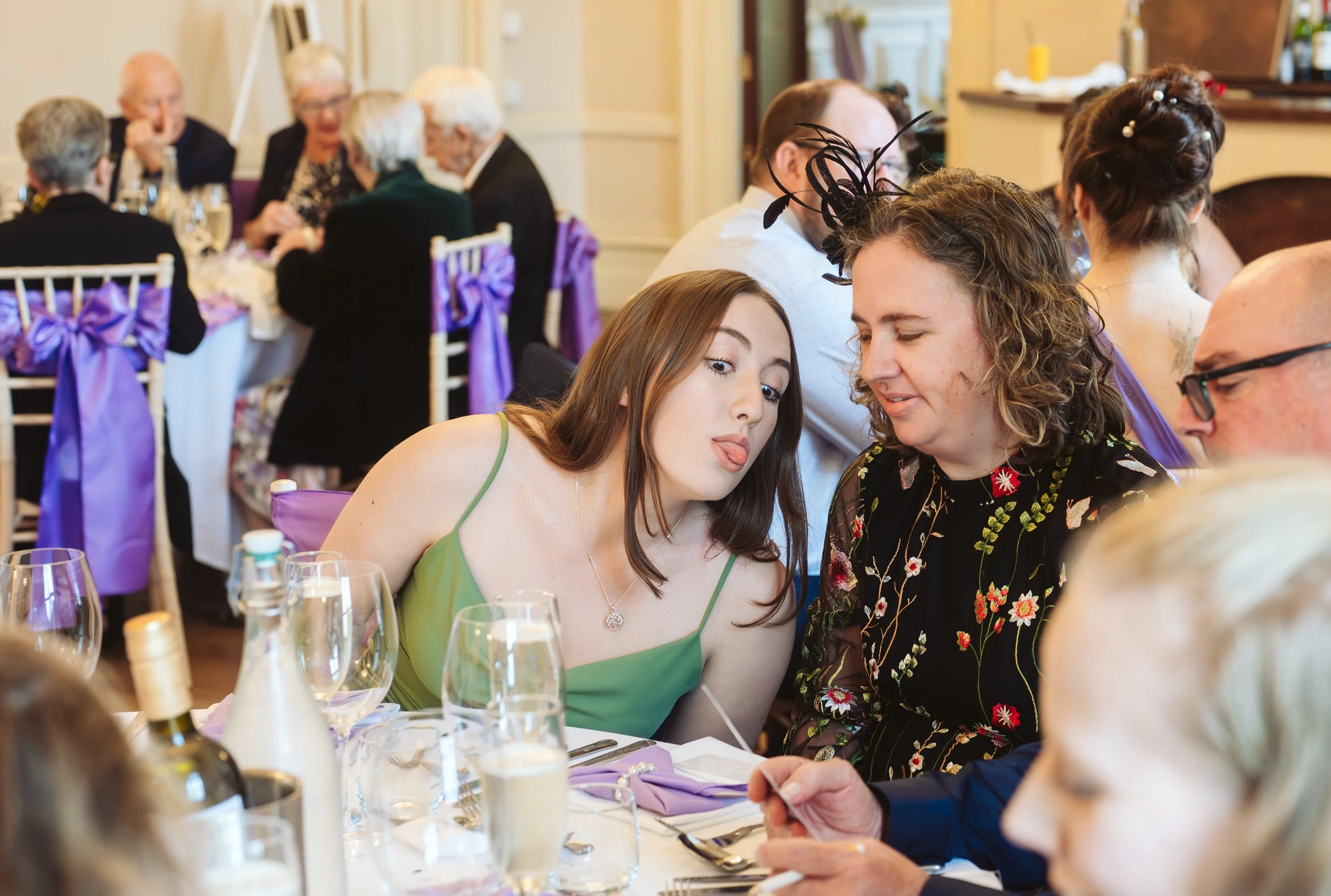 A young woman in a green dress making a funny face with her tongue out, sitting at a table with three or four older adults at a social gathering or wedding reception.