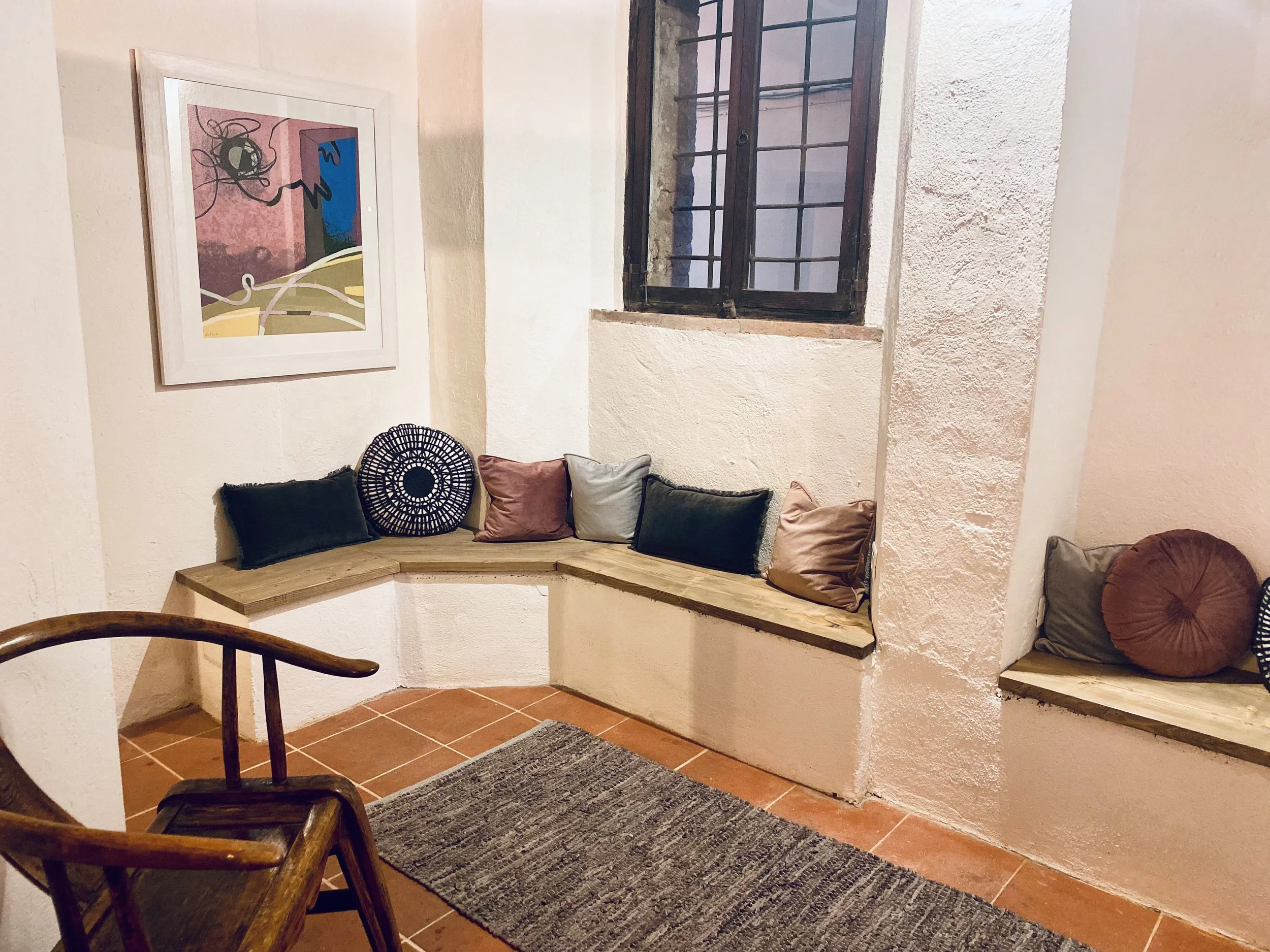 Cozy corner with built-in wooden bench seating adorned with multicolored pillows, a window with metal bars, a framed abstract art piece on the wall, a wooden chair, and a textured rug on terracotta tile floor.