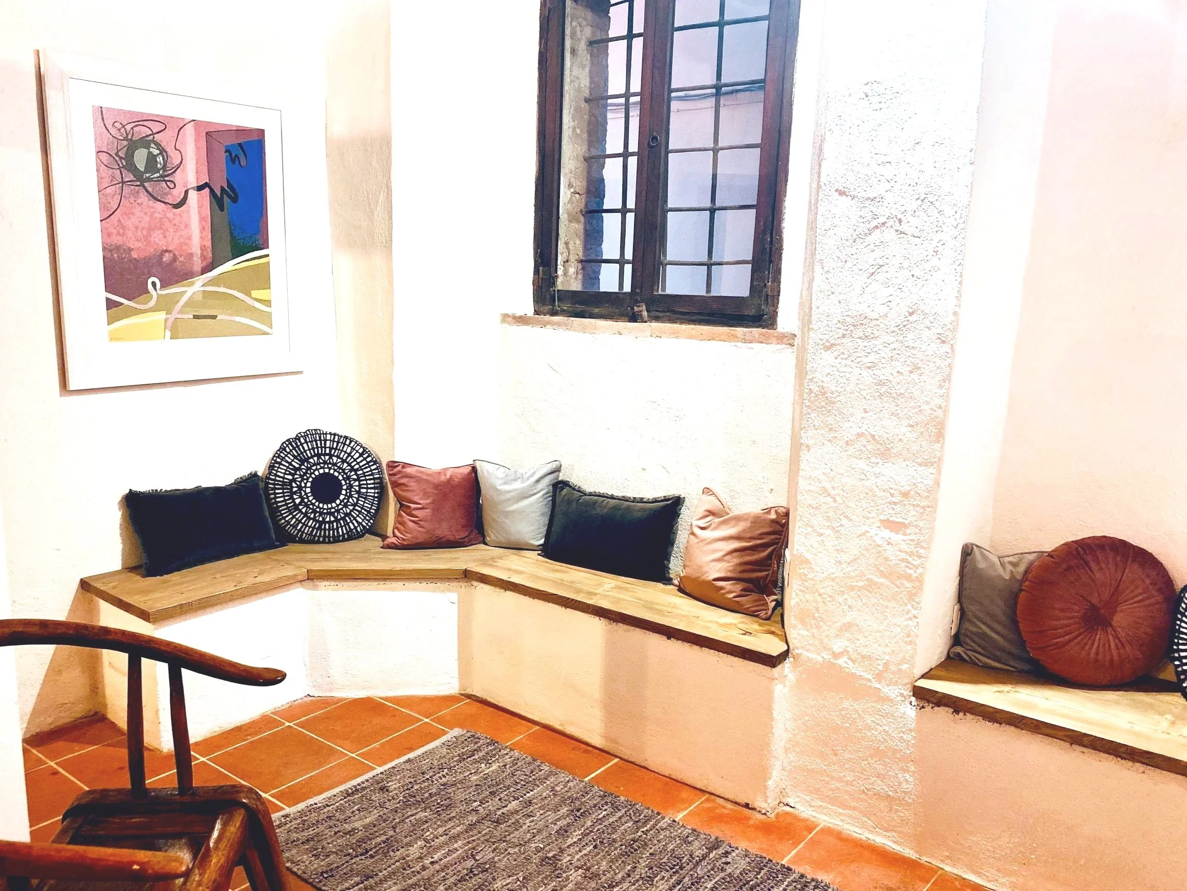 A cozy corner with a built-in wooden bench decorated with various colorful pillows in front of a textured white wall. To the left, a framed abstract artwork hangs on the wall. A small window with dark wooden framing and a grid of small windowpanes lets in natural light. The floor is tiled with reddish-brown tiles, and there is a dark woven rug near the bench.